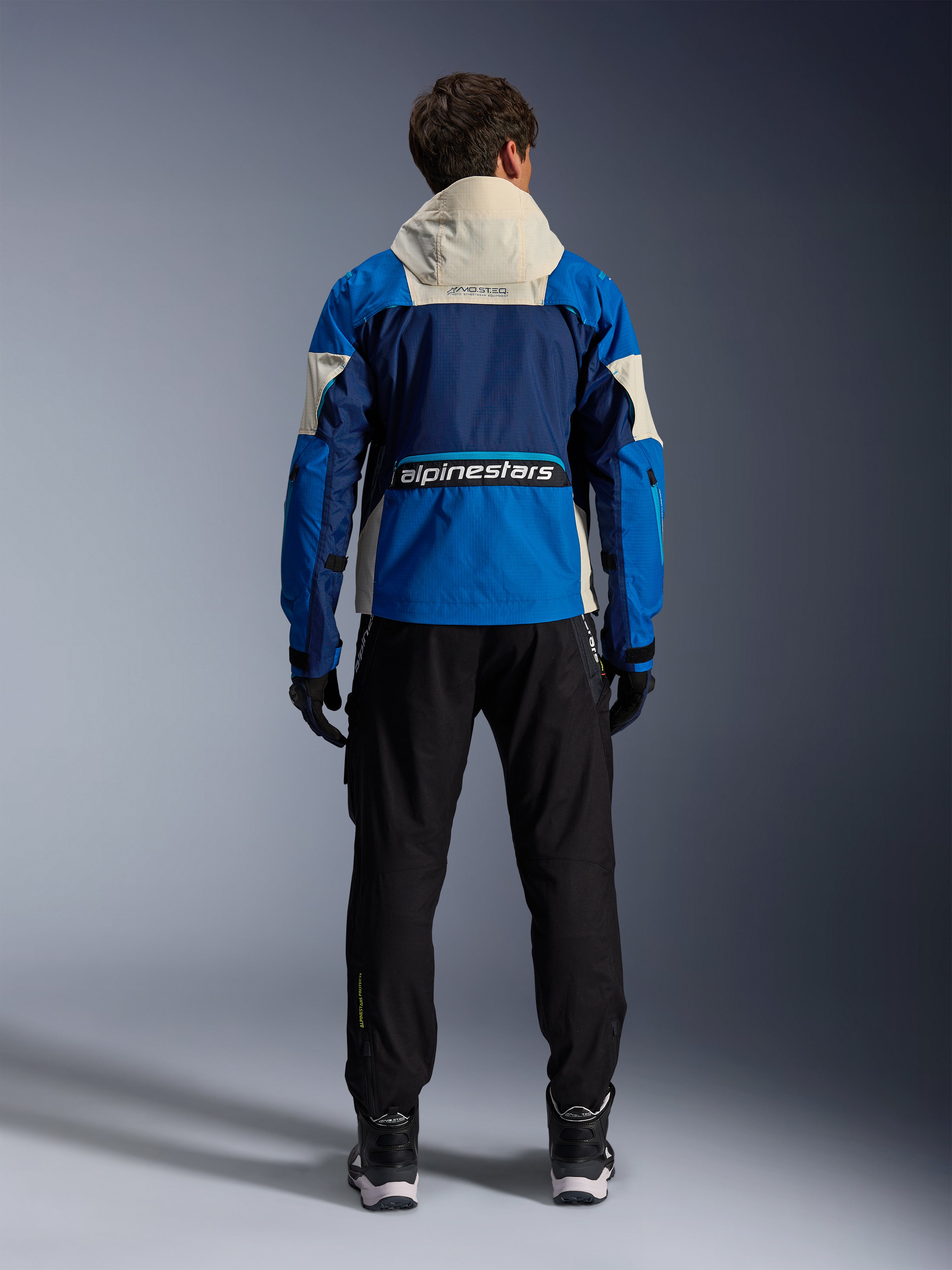 Alpinestars Mogress Air Jacket in Blue Almond, back view of a rider wearing the motorcycle jacket, featuring navy and bright blue panels with an off-white hood, bold Alpinestars branding on the lower back, urban contemporary riding gear style.