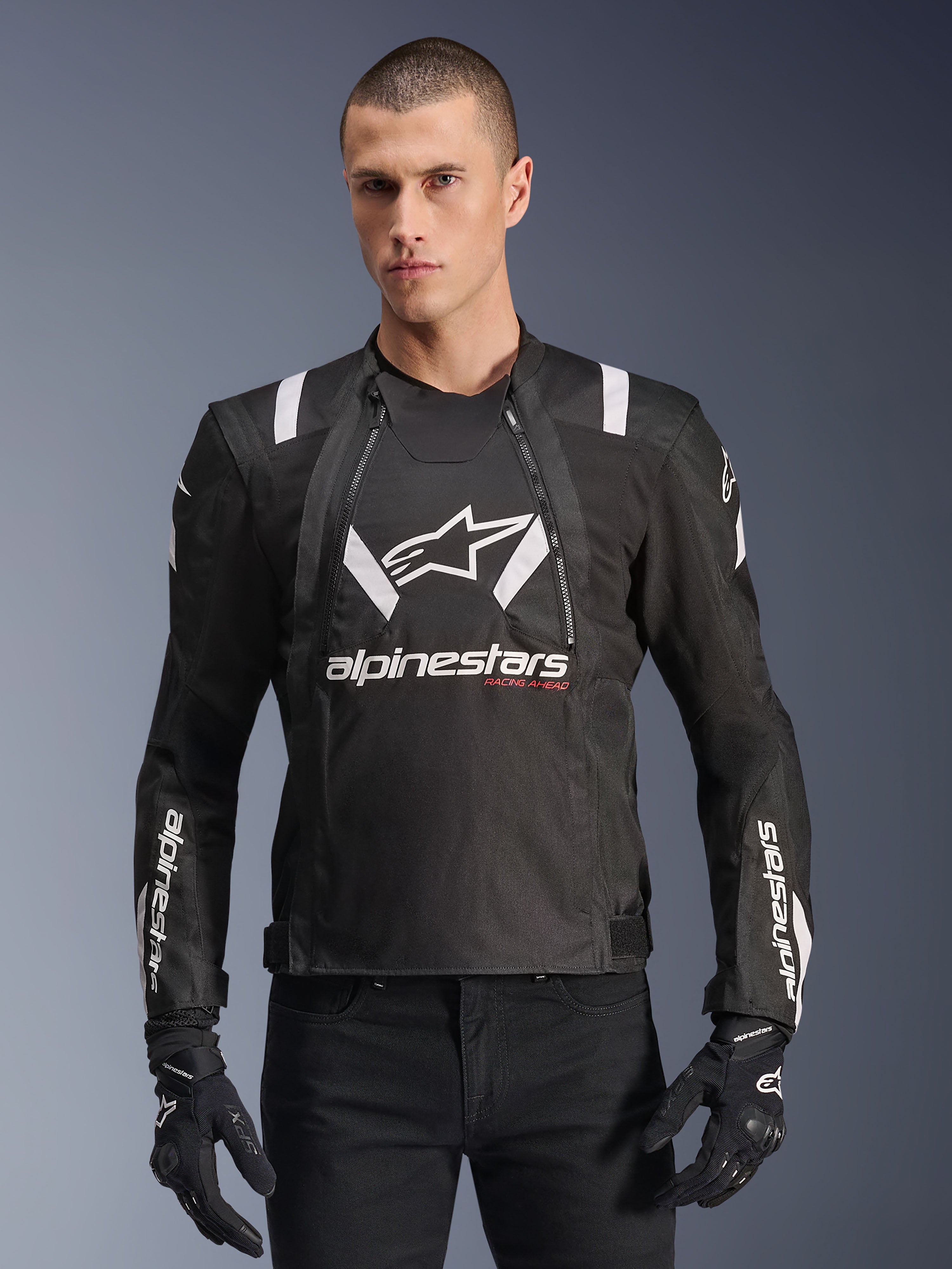 Alpinestars T-Stunt Air Jacket, svart och vit motorcykeljacka, bärs av en man med svarta handskar och byxor, framträdande vit logotyp på bröstet och ärmarna med röda 'Racing Ahead'-detaljer, sportig design med vita axelränder och ventilerande meshpaneler.