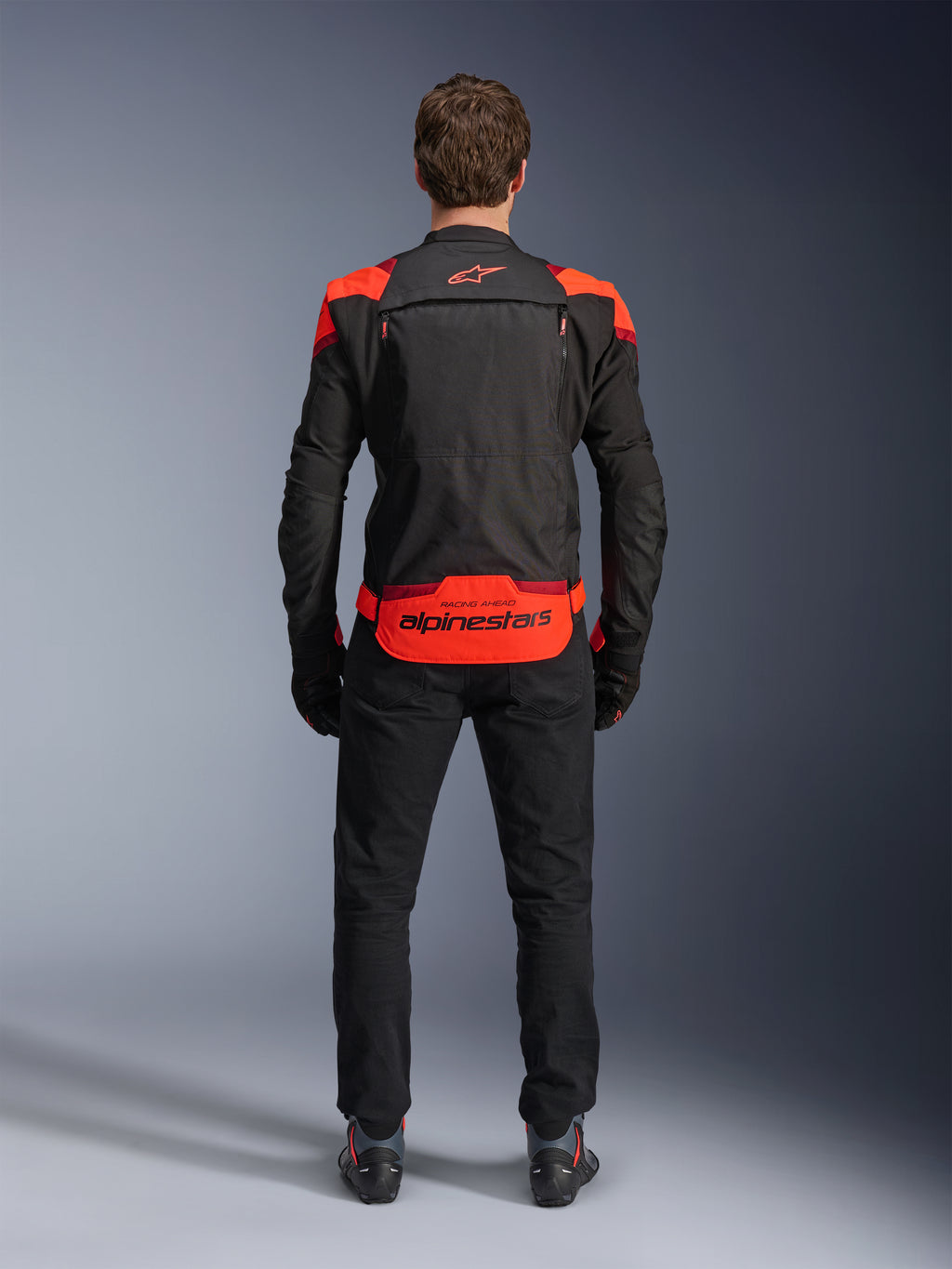 Alpinestars T-Stunt Air Jacket, Black with Red Fluo and Dark Red accents, rear view on model, paired with black riding pants and boots, featuring bold Alpinestars branding on the lower back panel, modular design with mesh construction for ventilation.