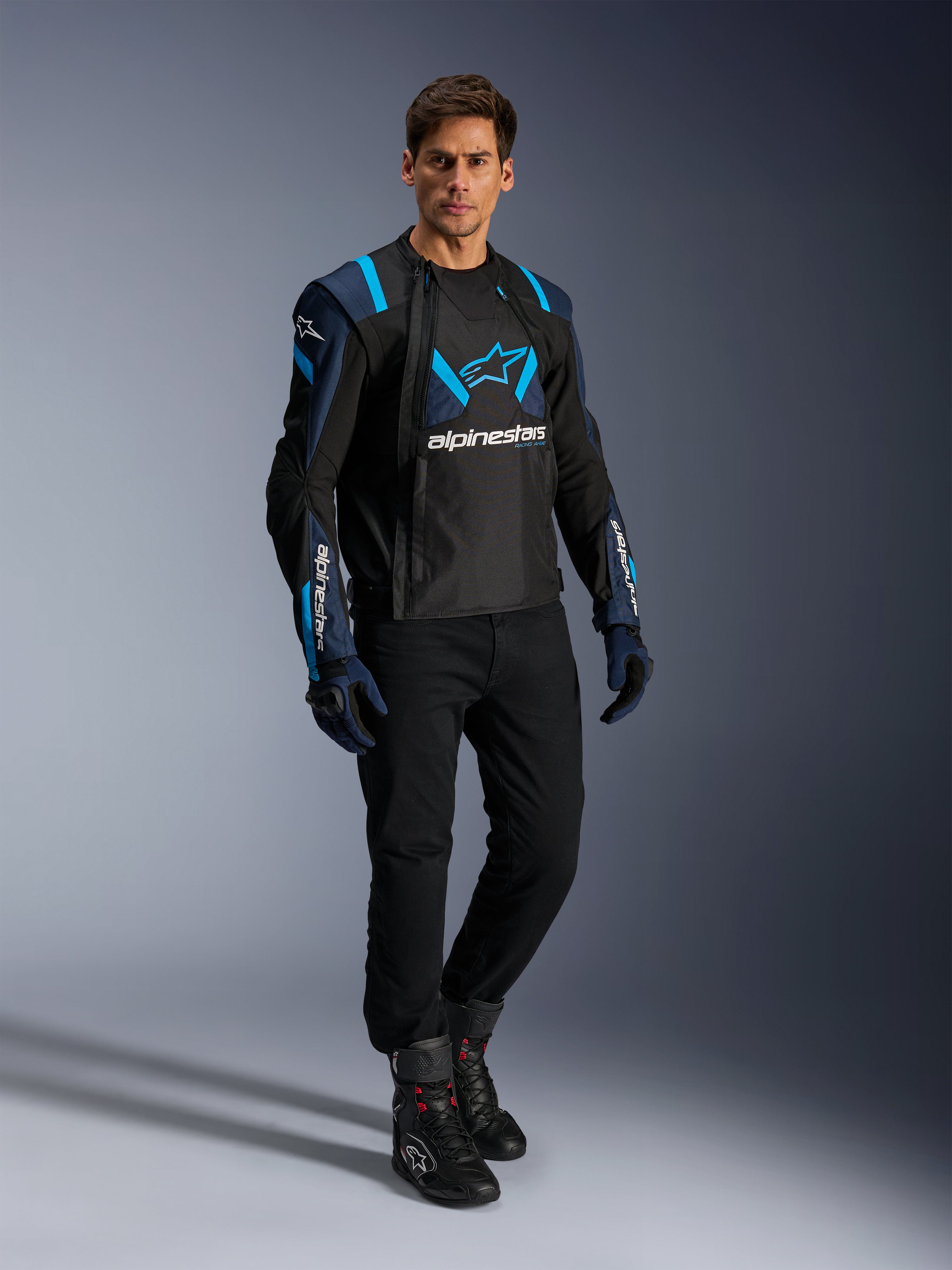 Manlig modell i Alpinestars T-Stunt Air-jacka, svart, mörkblå och Blithe Blue, med framträdande meshpaneler för luftflöde, matchad med svarta byxor och körhandskar, sportig motorcykeljacka för sommarkörning på väg.