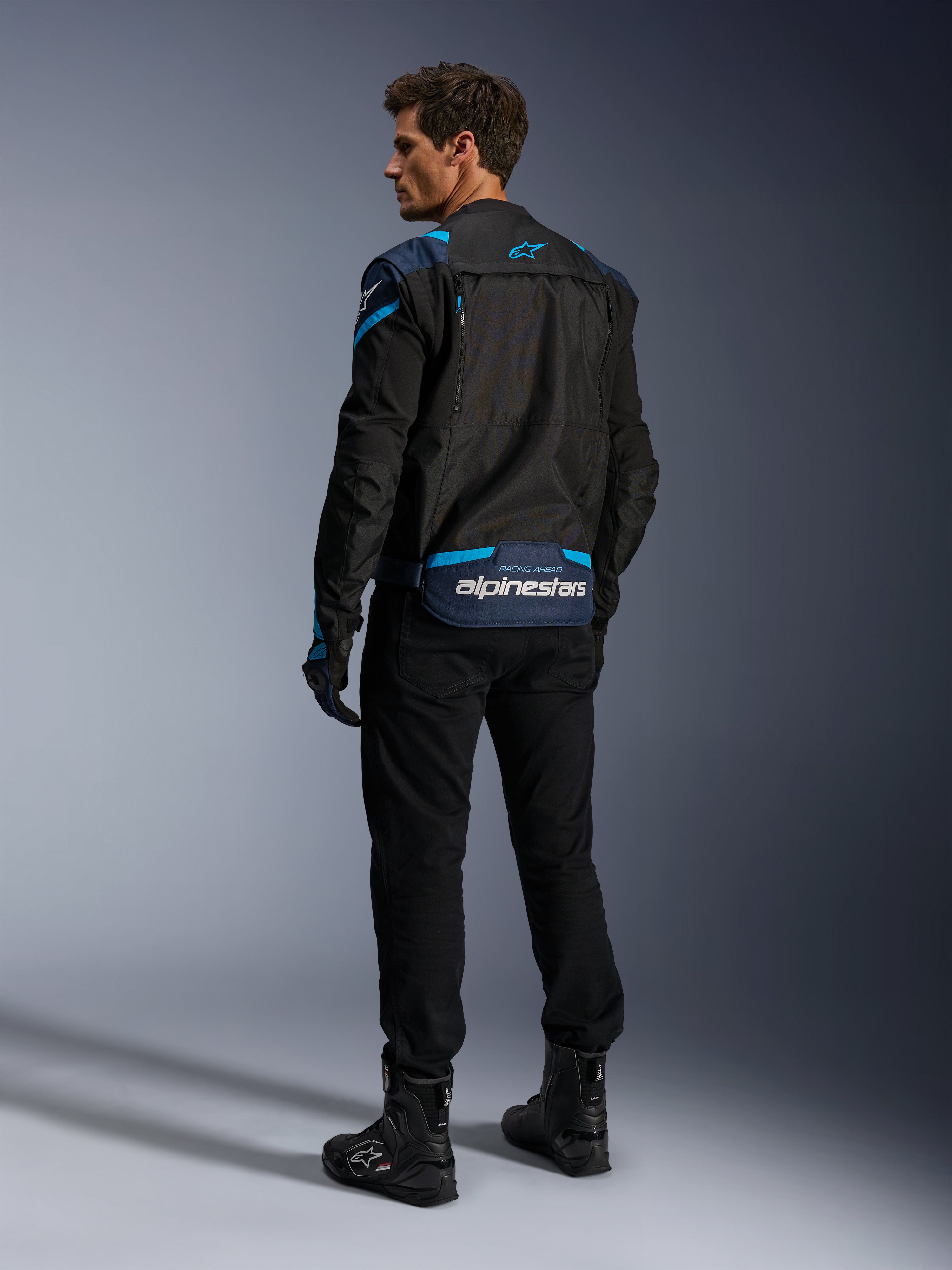 Alpinestars T-Stunt Air-jacka, motorcykeljacka, svart med mörkblå och ljusblå detaljer, vy bakifrån som visar modulära meshpaneler och logotyp på nedre delen av ryggen, bärs av en modell med svarta körbyxor och stövlar.