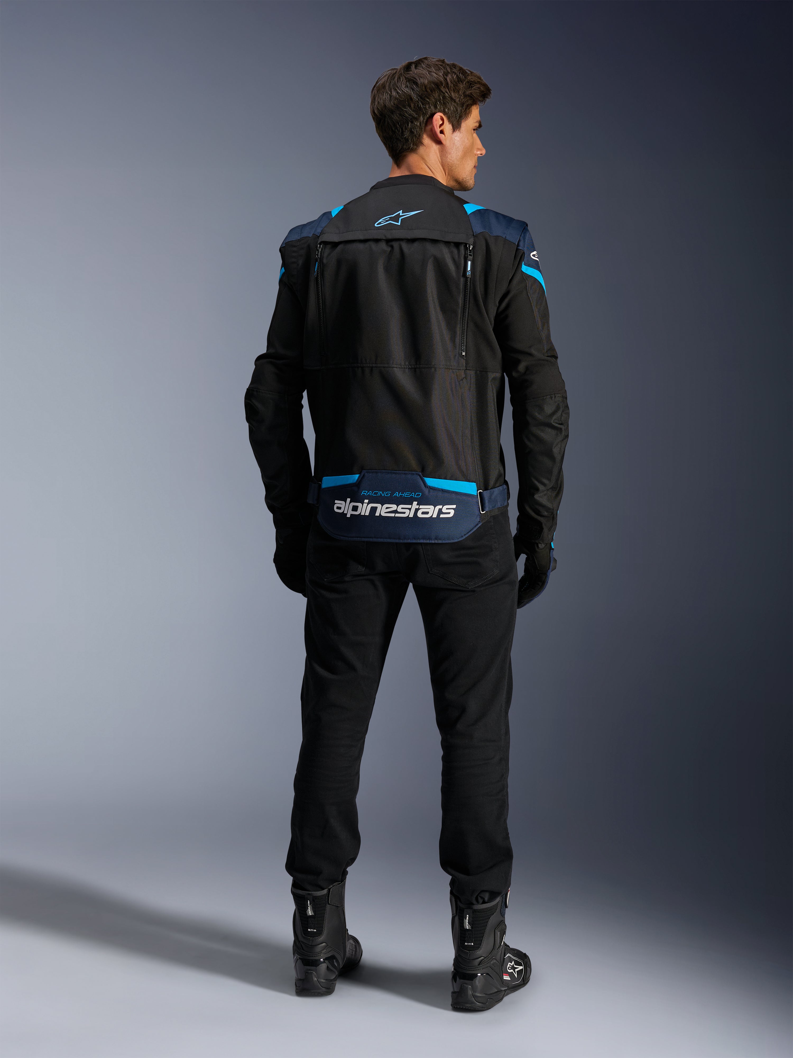 Alpinestars T-Stunt Air-jacka, modulär textiljacka för motorcykel, svart, mörkblå och ljusblå, ryggvy som visar det nedre ryggfacket för förvaring av ärmar med vit Alpinestars-logotyp, bärs med svarta byxor och stövlar.