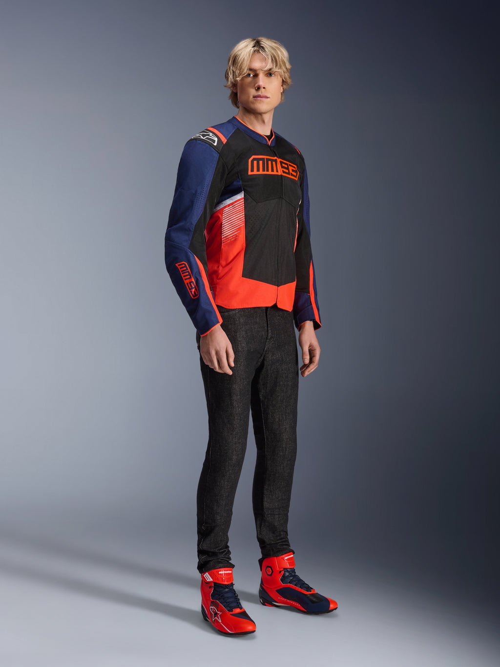 Manlig modell iklädd Alpinestars MM93 T-SPS Air V2-jackan i Blue Navy, svart och Red Fluo, med framträdande MM93-logotyp på bröstet och ärmarna, kombinerad med mörka jeans och matchande röda och blå motorcykelstövlar, sportig urban mc-utrustning
