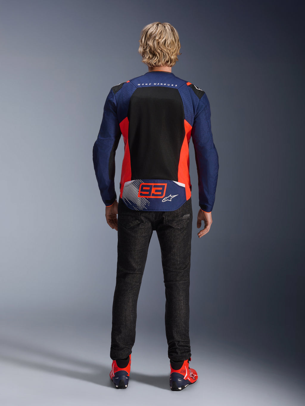 Ryggvy av en modell iklädd MM93 T-SPS Air V2-jacka i marinblått med Red Fluo och svarta detaljer, med en stor meshpanel på ryggen och Marc Marquez '93'-branding, kombinerat med mörka denimbyxor och matchande mc-stövlar