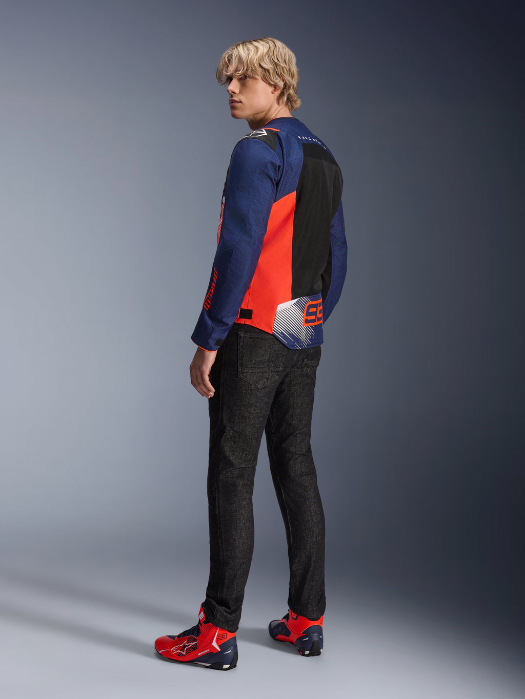 Alpinestars MM93 T-SPS Air V2-jacka, Blue Navy och röd fluo, bärs av en modell bakifrån med svarta mc-jeans och matchande röda och blå skor, med meshpaneler och Marc Márquez 93-logotyp.