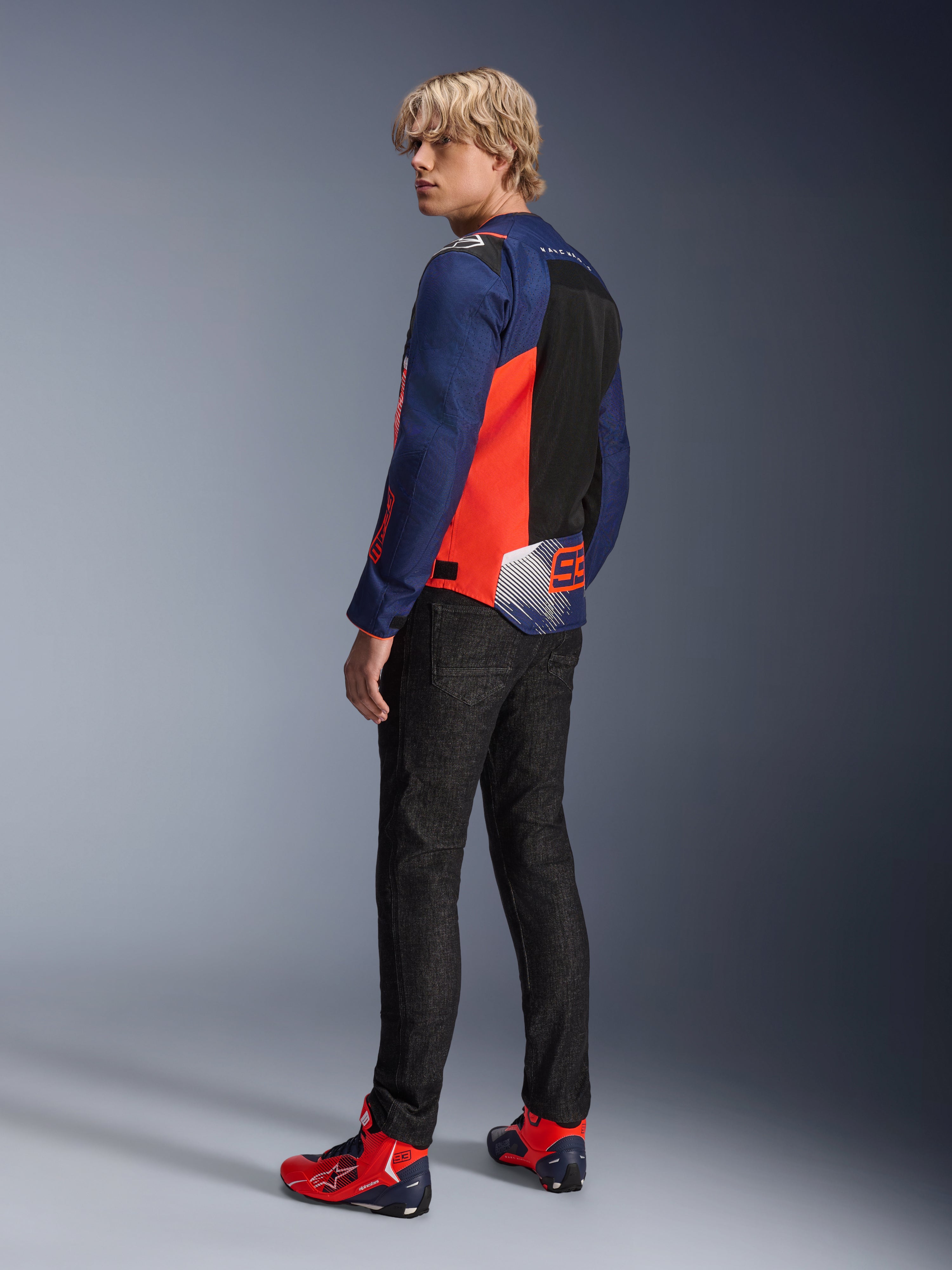 Alpinestars MM93 T-SPS Air V2-jacka, Blue Navy och röd fluo, bärs av en modell bakifrån med svarta mc-jeans och matchande röda och blå skor, med meshpaneler och Marc Márquez 93-logotyp.