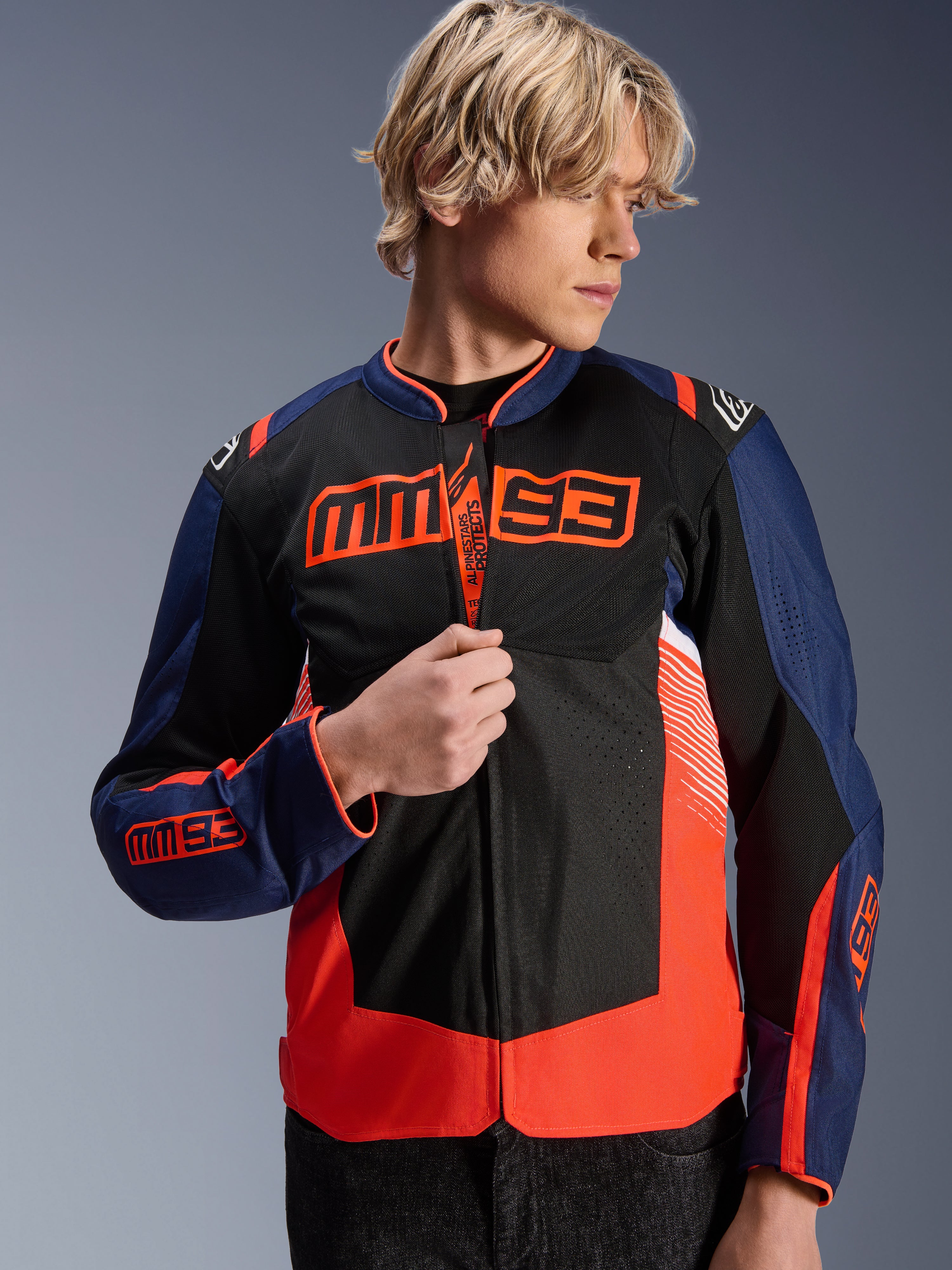 Alpinestars MM93 T-SPS Air V2-jacka, mörkblå, svart och fluorescerande röd motorcykeljacka i textil, bärs av en manlig modell som justerar dragkedjan, med framträdande Marc Márquez MM93-logotyp på bröstet och ärmarna, med ventilerande meshpaneler för sommarkörning.