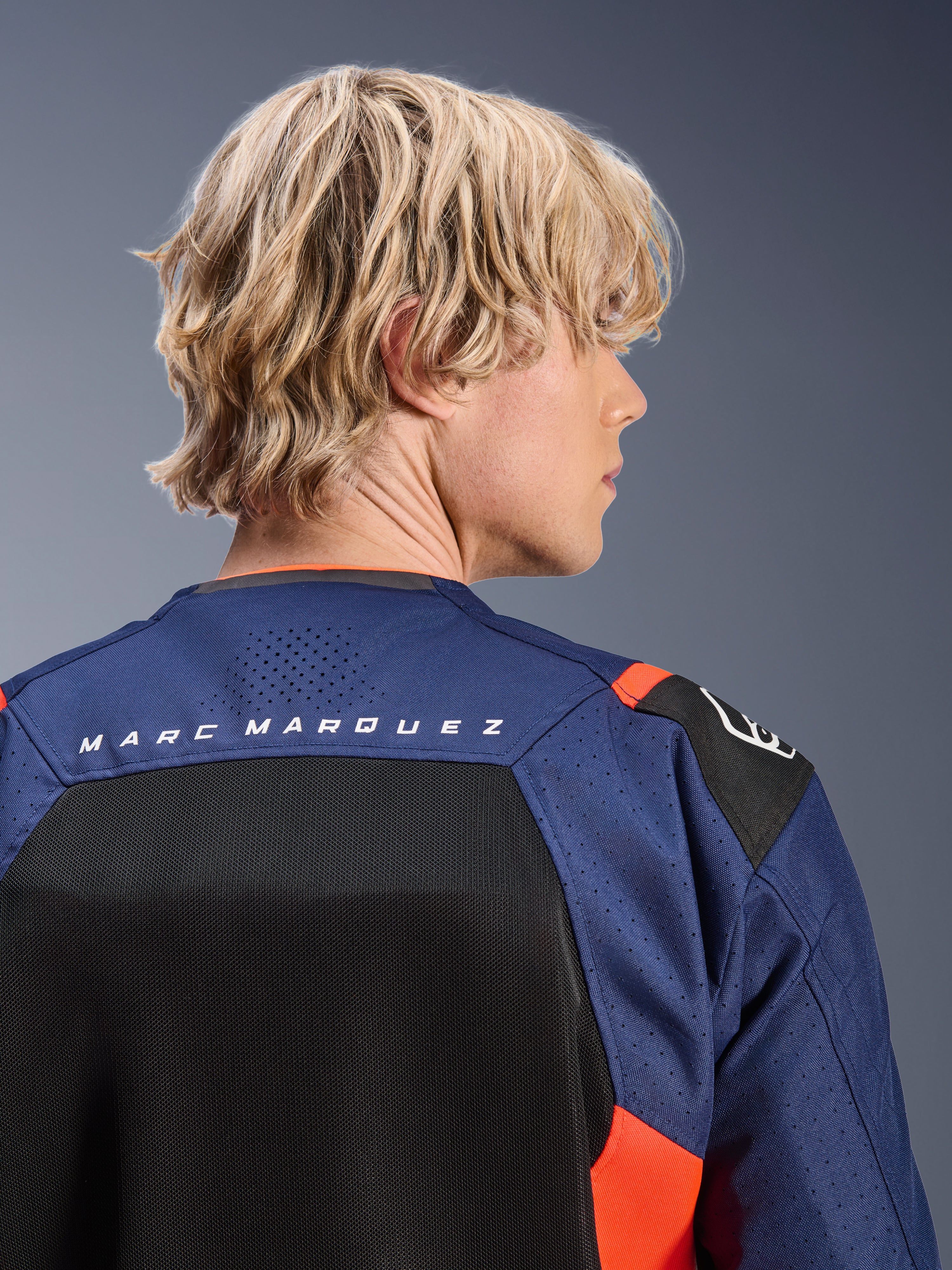 Alpinestars MM93 T-SPS Air V2-jacka, mörkblå och svart med röda fluo-detaljer, närbild på övre ryggen med 'MARC MARQUEZ'-logotyp, ventilerande meshpaneler och perforerad textilkonstruktion, bärs av en blond modell.