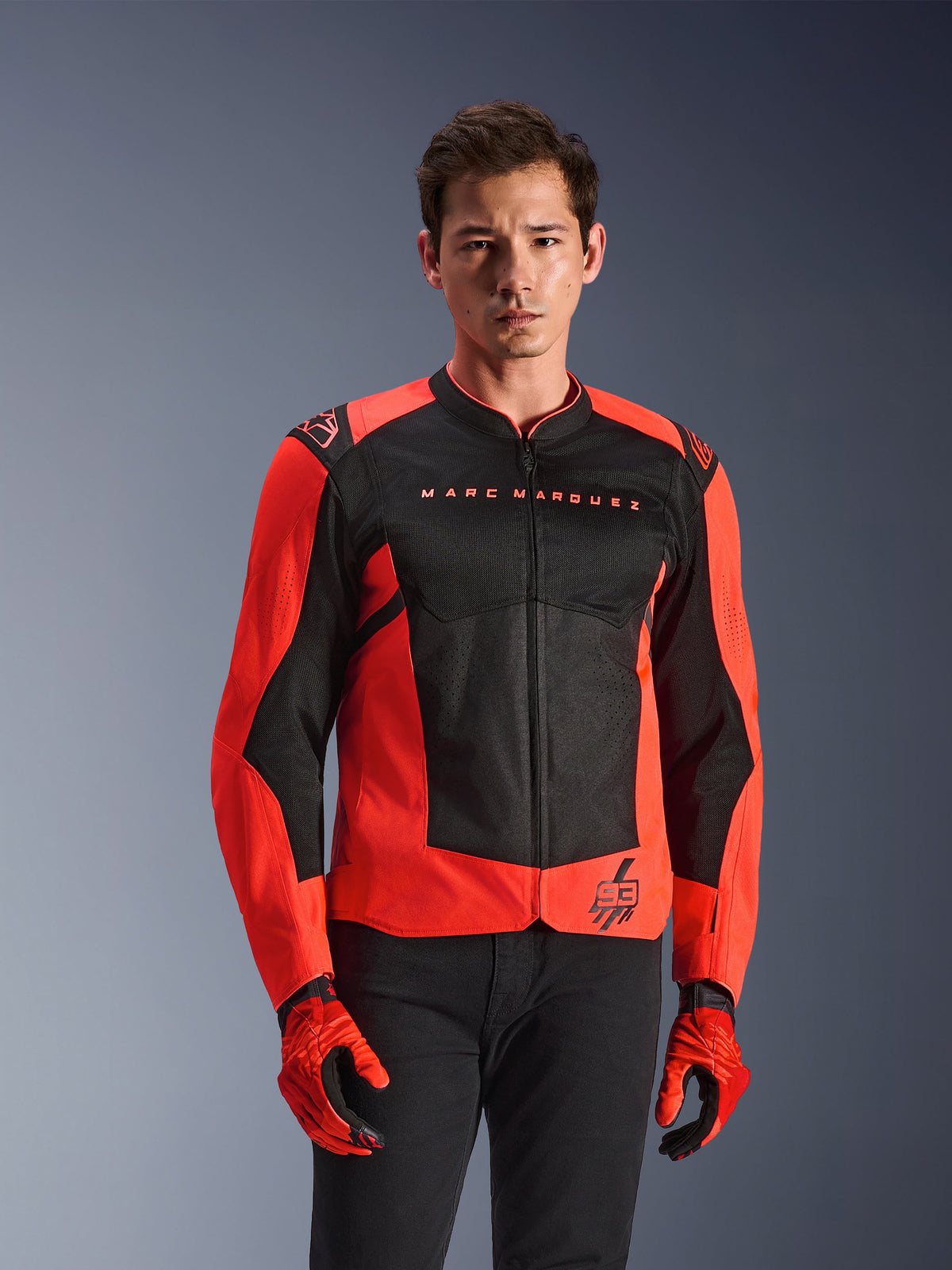 Alpinestars MM93 T-SPS Air V2-jacka, motorcykeljacka, röd fluo och svart, bärs av en modell med matchande röda handskar och svarta byxor, med 'Marc Marquez'-märkning på bröstet och '93'-logotyp, sportig mesh-konstruktion