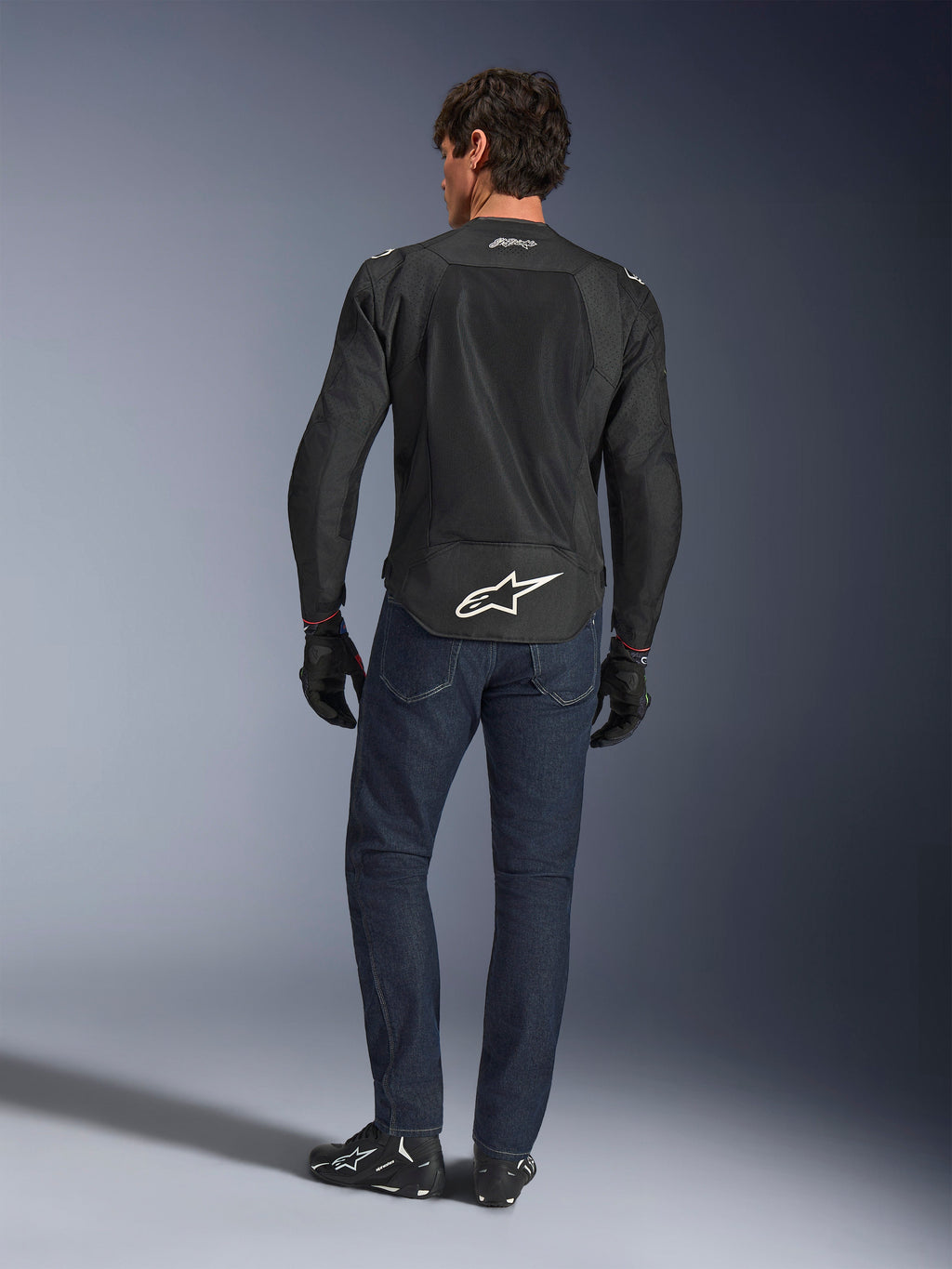 Alpinestars FQ20 T-SPS Air V2 Monster Jacka, svart, vy bakifrån på modell i mörkblå jeans och svarta MC-stövlar, stor vit logotyp på nedre delen av ryggen och ventilerande meshpaneler, urban sportig MC-utrustning