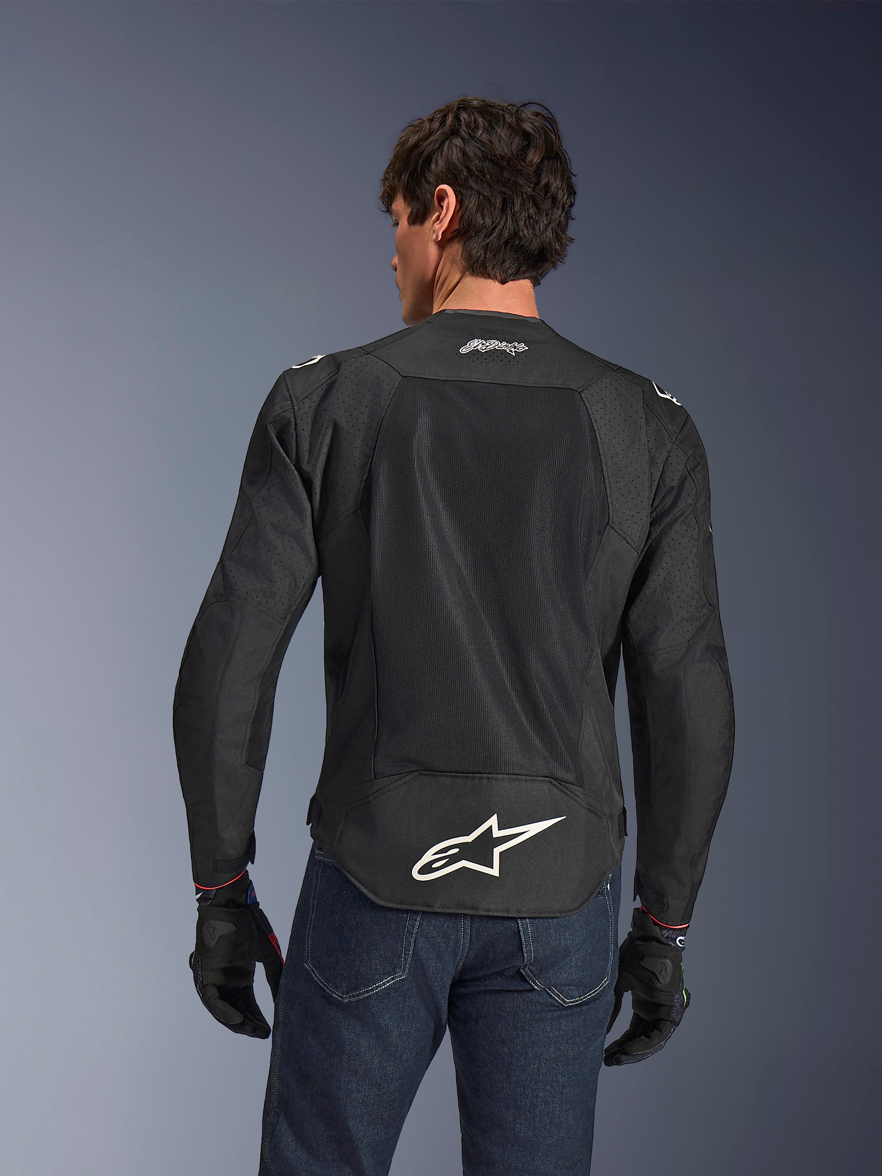 Alpinestars FQ20 T-SPS Air V2 Monster-jacka, svart, vy bakifrån med stora ventilationspaneler i mesh och vit Alpinestars-logotyp på nedre delen av ryggen, bärs med blå jeans och svarta motorcykelhandskar, urban sportig körstil.