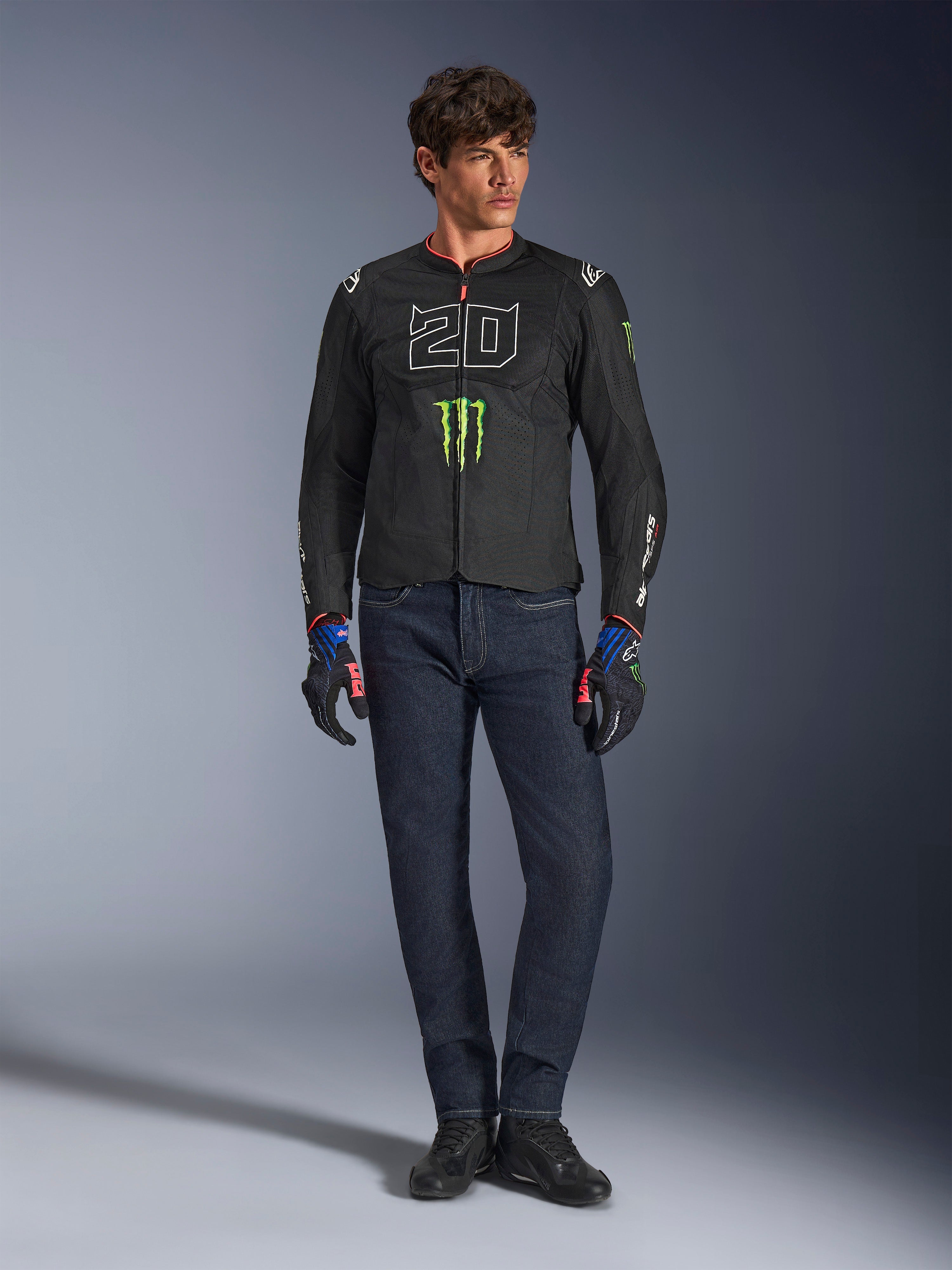 Manlig förare bärande Alpinestars FQ20 T-SPS Air V2 Monster-jacka, svart med grön Monster Energy-logotyp och vit '20'-grafik på bröstet, kombinerat med mörkblå jeans och tekniska körhandskar, urban sportig motorcykeloutfit.