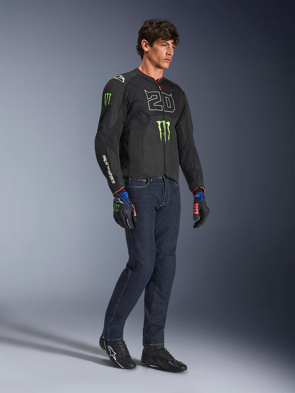 Manlig förare som bär Alpinestars FQ20 T-SPS Air V2 Monster-jacka i svart, med '20'-logotypen och Monster Energy-klon på bröstet, kombinerat med mörka MC-jeans, svarta MC-skor och matchande FQ20-handskar, studiobild i helfigur.