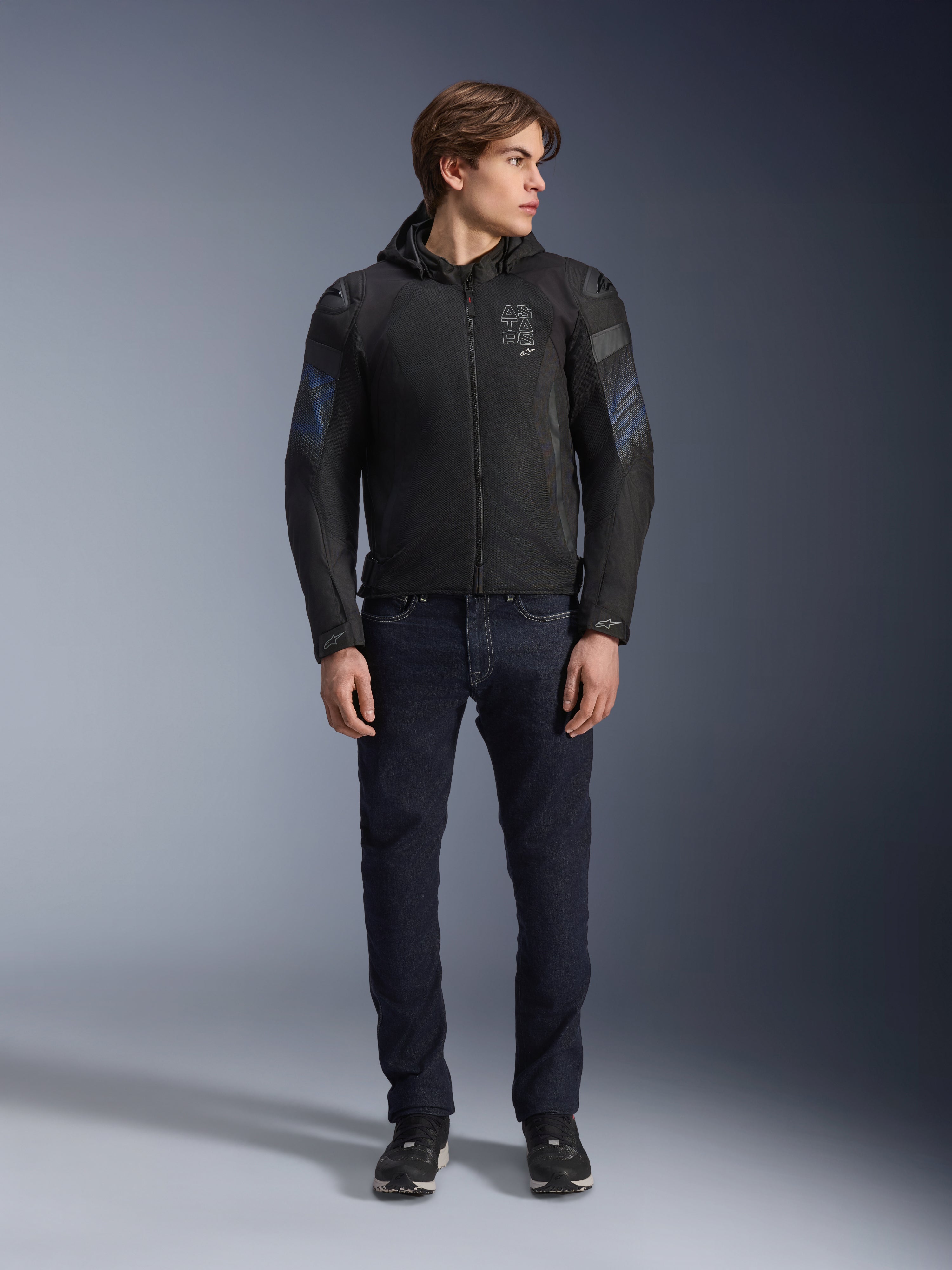 Male model wearing Alpinestars Zaca Air Venom WP Jacket in Black and Electric Blue, textile motorcycle jacket with 3D mesh panels and a detachable hood, paired with dark denim jeans, modern sport-inspired riding gear