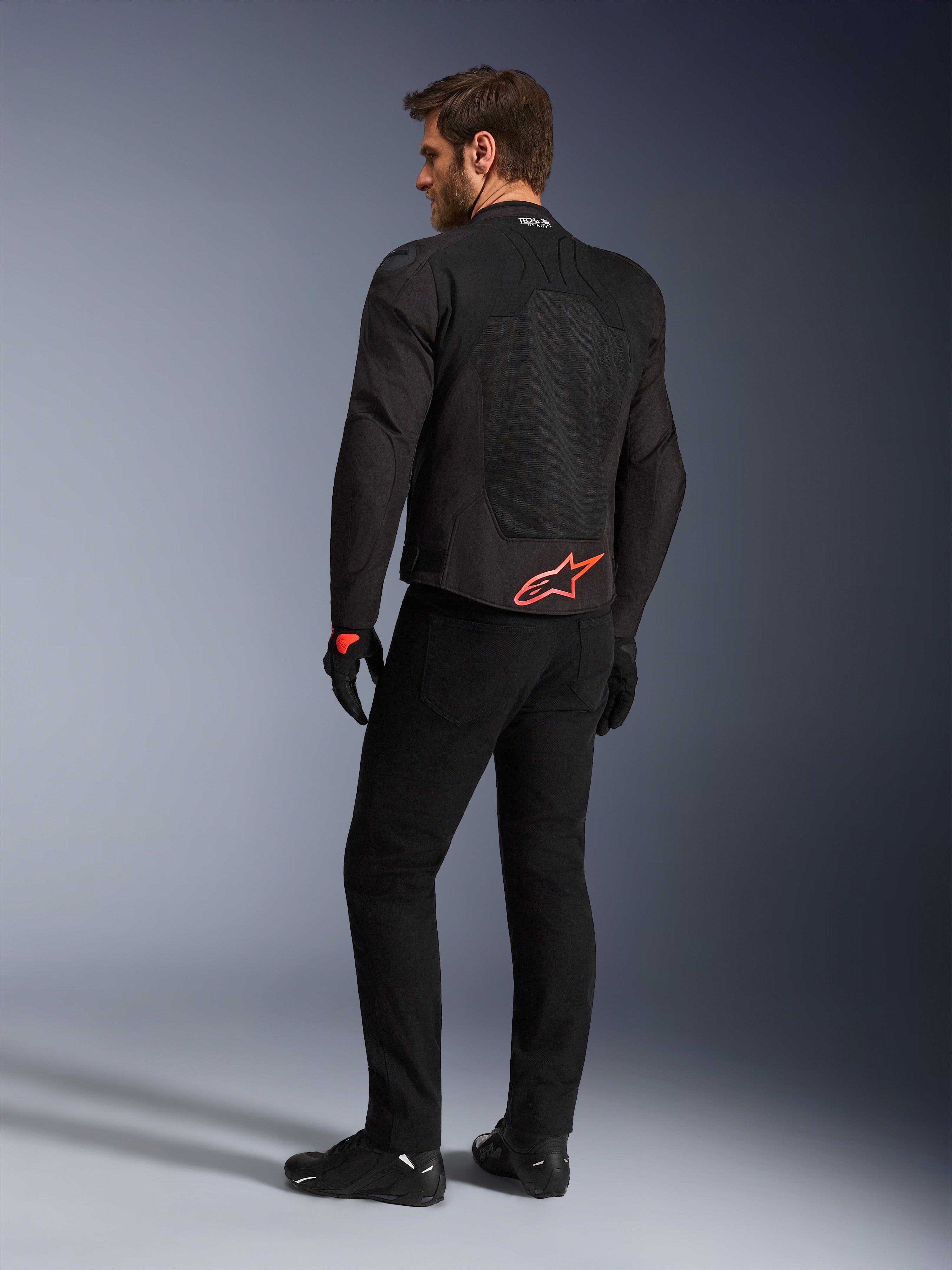 Bakifrån vy av en modell som bär Alpinestars SMX Air Jacket i svart, med en klaröd Alpinestars-logotyp på nedre delen av ryggen, 'Tech-Air Ready'-märkning på kragen och tekniska mesh-paneler för ventilation, matchad med svarta körbyxor och handskar.