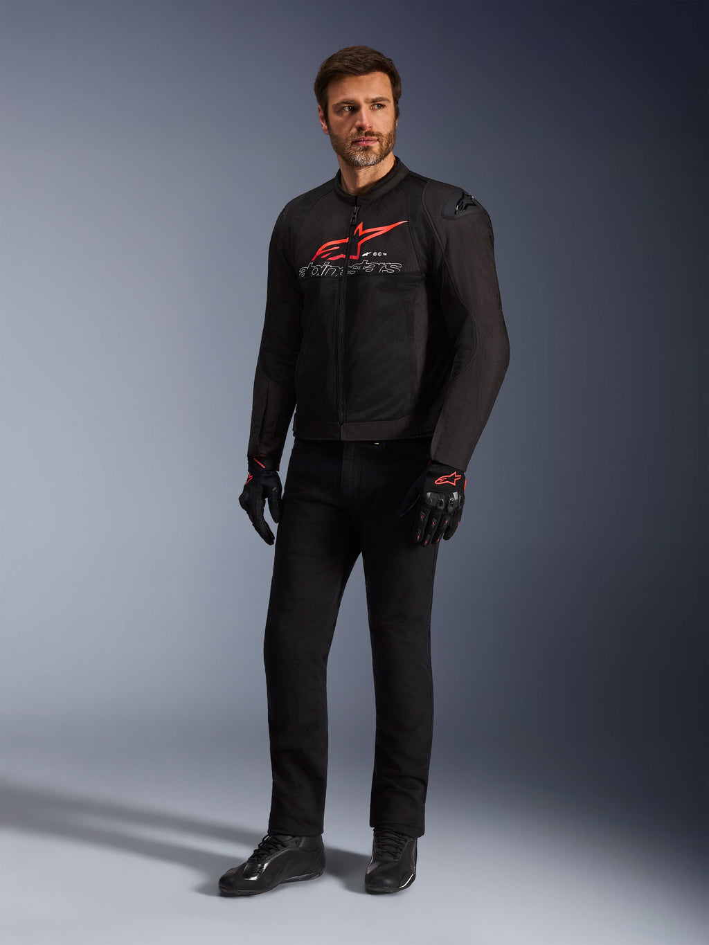 Manlig modell som bär Alpinestars SMX Air-jacka i svart, klarrött och vitt, stylad med svarta tekniska byxor, svarta och röda handskar och svarta mc-skor, med en framträdande röd logotyp på bröstet och vit branding, helkroppsbild i studiomiljö för sportmotorcykelutrustning