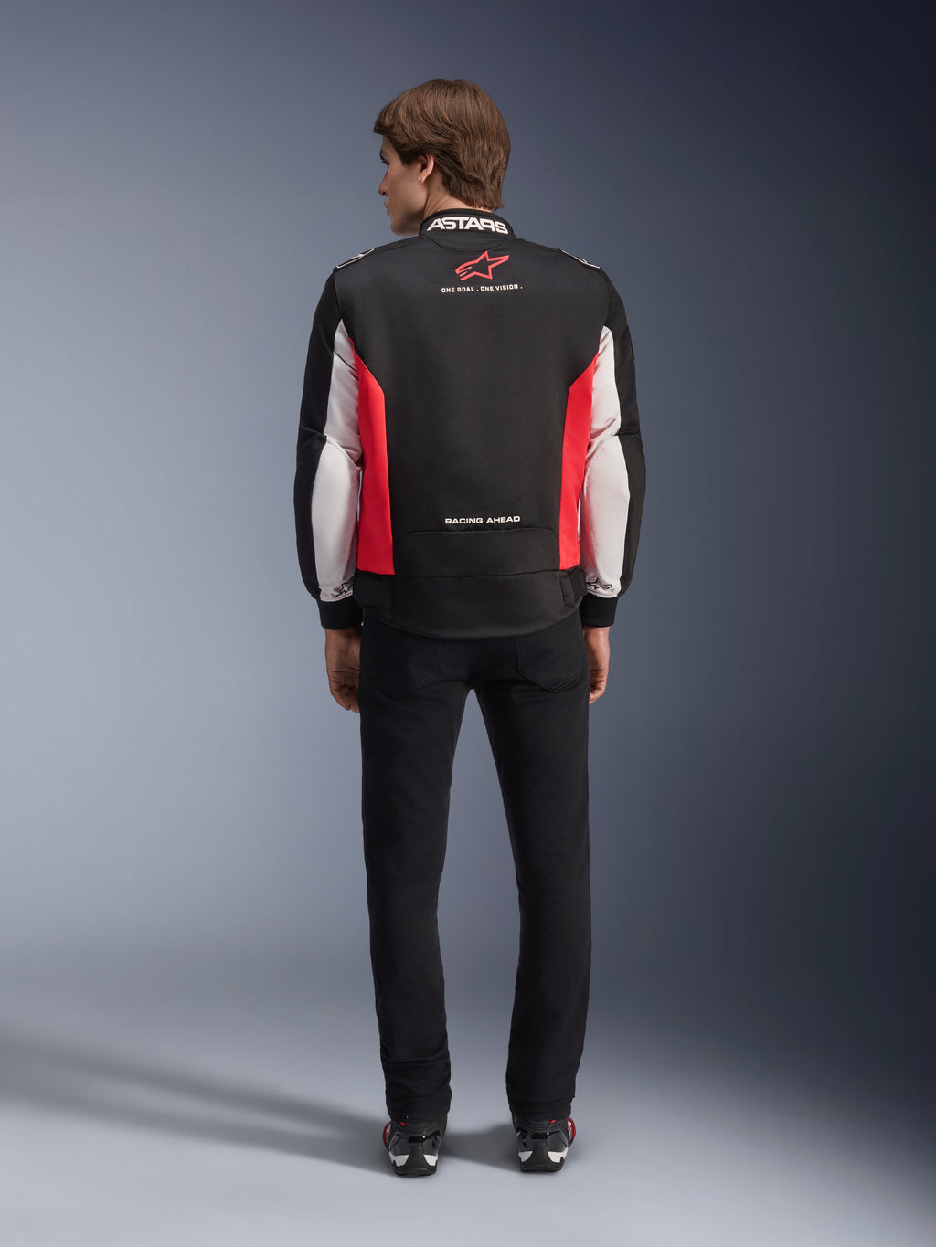 Alpinestars Monza Sport Jacket, svart, klarröd och vit textiljacka för motorcykel, sedd bakifrån på en modell, med röd stjärnlogotyp och 'ASTARS'-märkning på kragen samt slogans, bärs med svarta byxor och körstövlar.