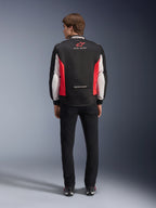 Alpinestars Monza Sport Jacket, svart, klarröd och vit textiljacka för motorcykel, sedd bakifrån på en modell, med röd stjärnlogotyp och 'ASTARS'-märkning på kragen samt slogans, bärs med svarta byxor och körstövlar.