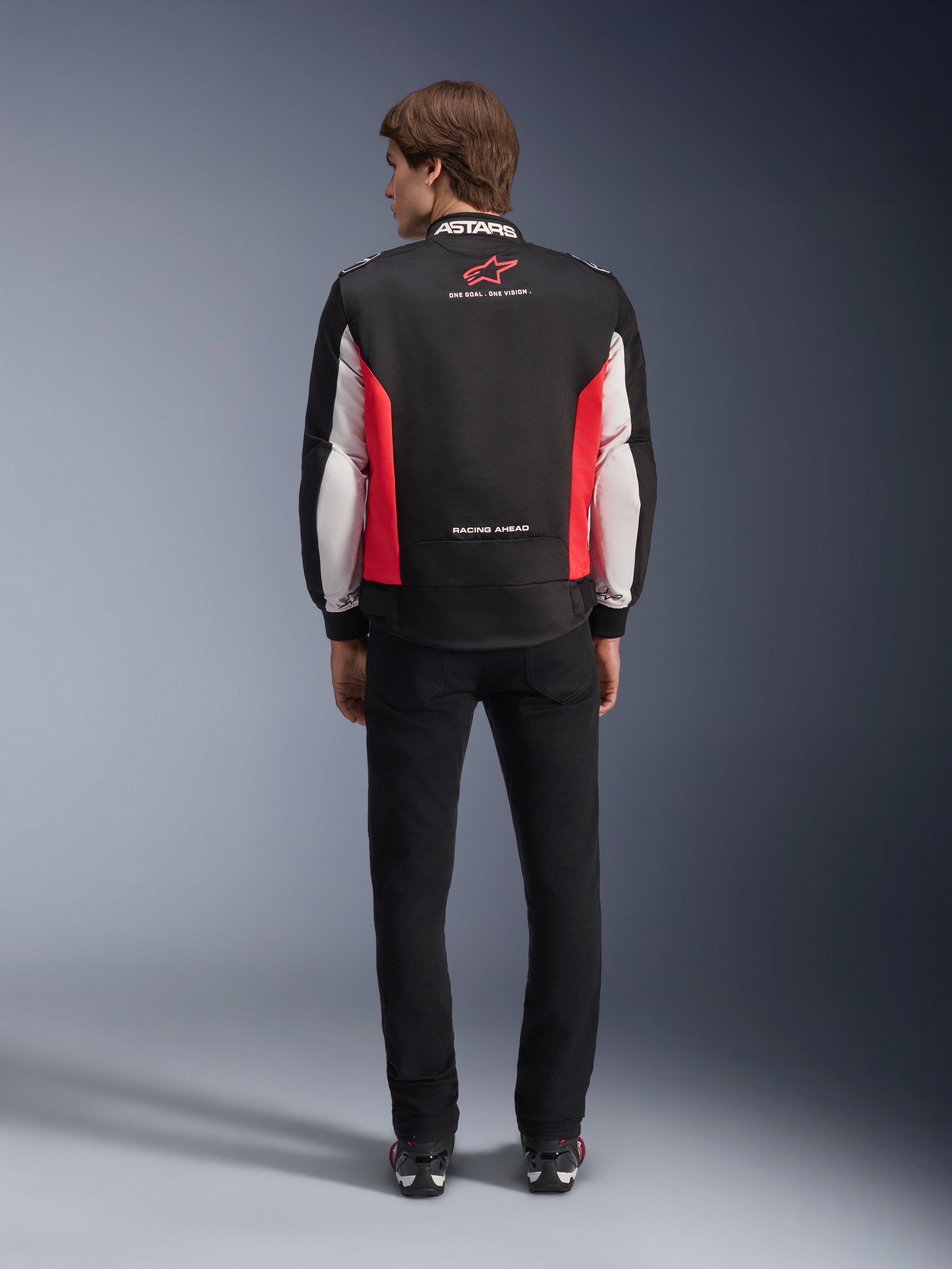 Alpinestars Monza Sport Jacket, svart, klarröd och vit textiljacka för motorcykel, sedd bakifrån på en modell, med röd stjärnlogotyp och 'ASTARS'-märkning på kragen samt slogans, bärs med svarta byxor och körstövlar.