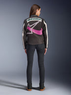 Bakifrån av en kvinna som bär Alpinestars Idles Jacket, svart med off-white ränder, med en stor lila och vit Alpinestars stjärnlogotyp och grön ordlogotyp på ryggen, kombinerad med mörka slim-fit jeans, vintageinspirerade urbana motorcykelkläder