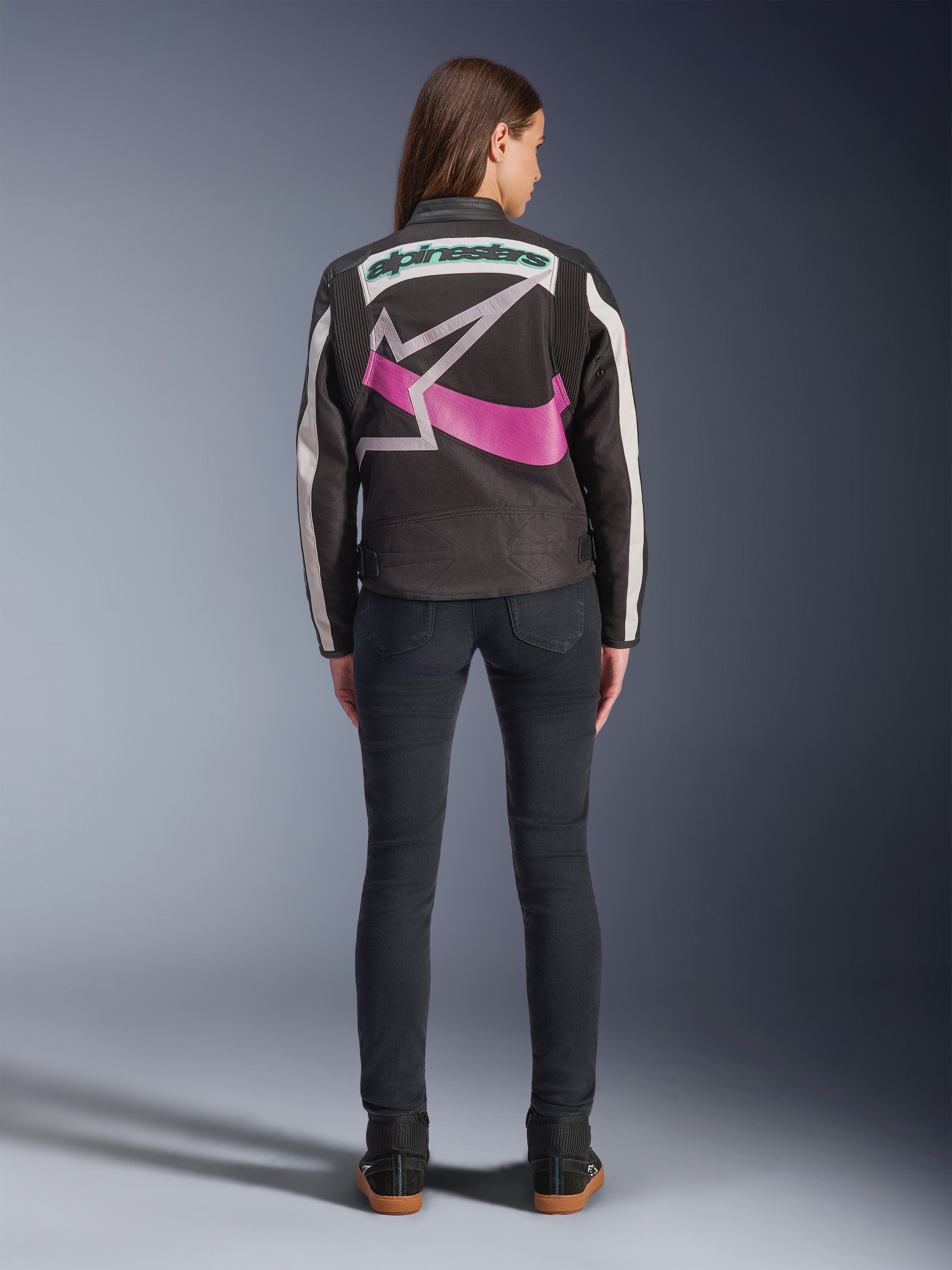 Bakifrån av en kvinna som bär Alpinestars Idles Jacket, svart med off-white ränder, med en stor lila och vit Alpinestars stjärnlogotyp och grön ordlogotyp på ryggen, kombinerad med mörka slim-fit jeans, vintageinspirerade urbana motorcykelkläder
