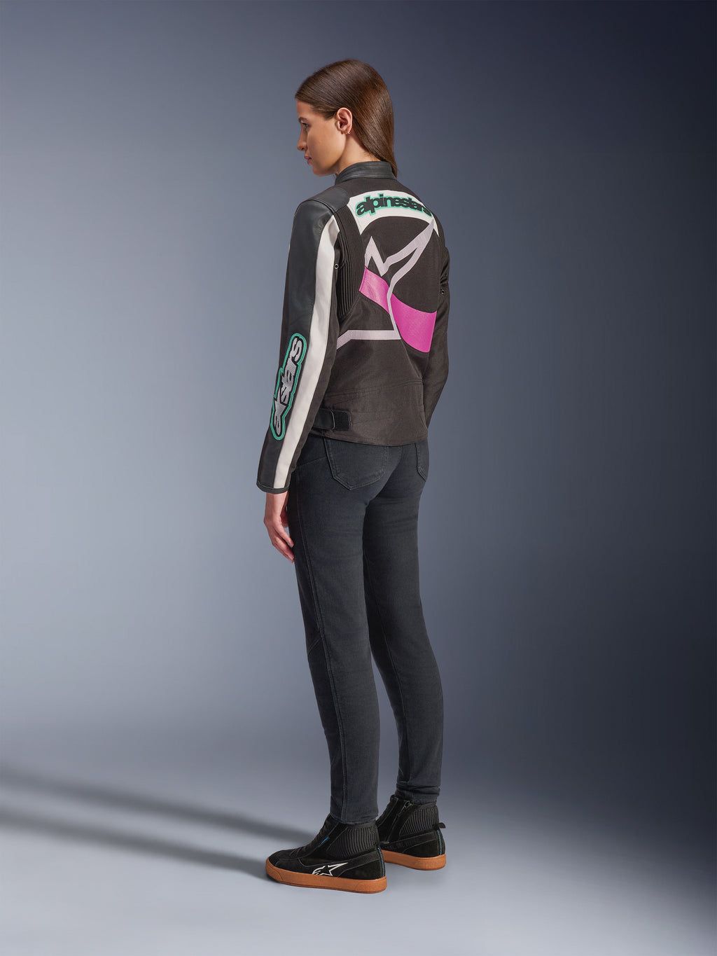 Alpinestars Idles damjacka, motorcykeljacka, svart, off-white och lila, sedd bakifrån på modell, med en stor omslutande Alpinestars-logotyp och vintage racing-inspirerad design, matchad med mörka jeans och svarta skor