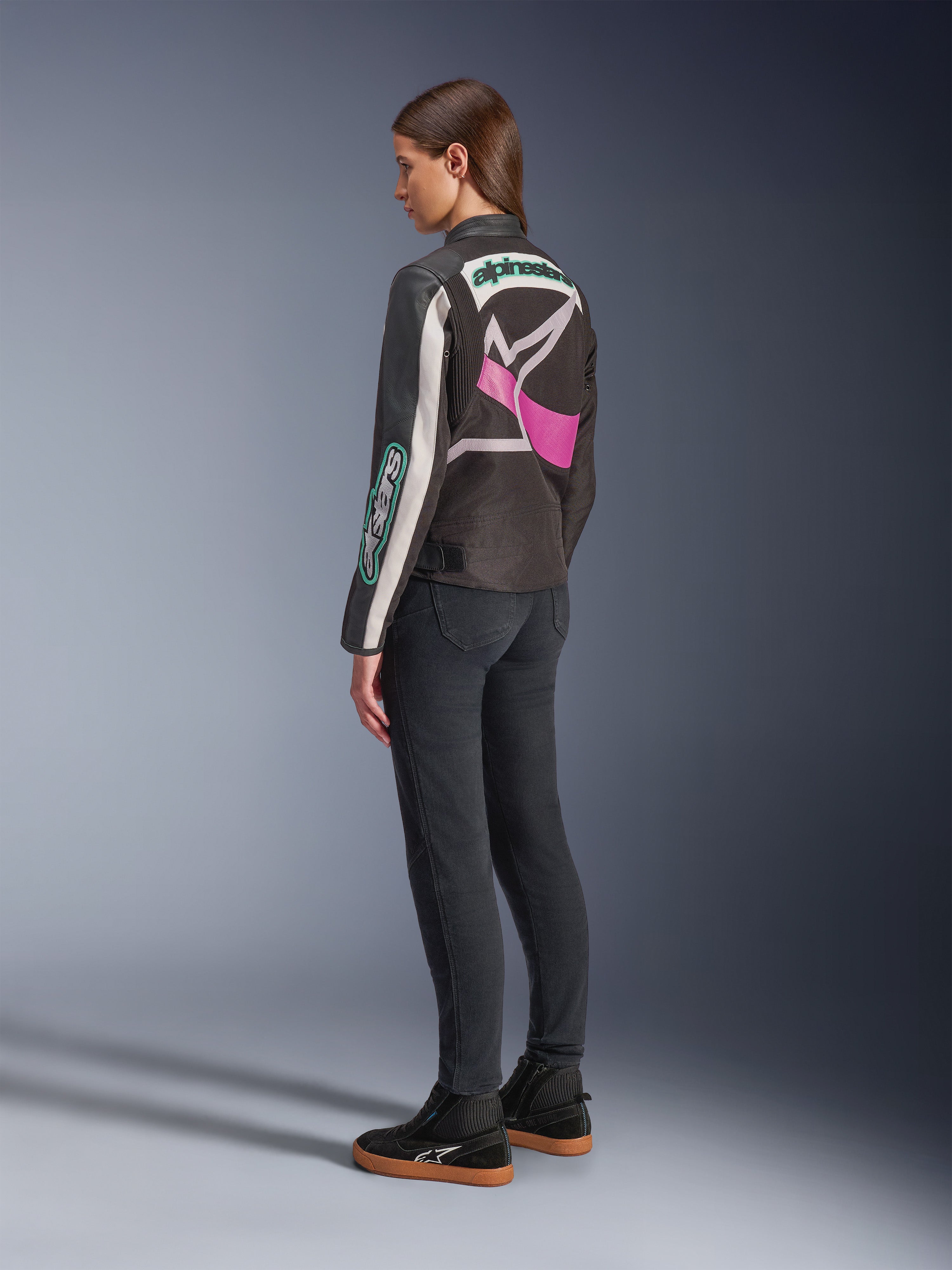 Alpinestars Idles damjacka, motorcykeljacka, svart, off-white och lila, sedd bakifrån på modell, med en stor omslutande Alpinestars-logotyp och vintage racing-inspirerad design, matchad med mörka jeans och svarta skor