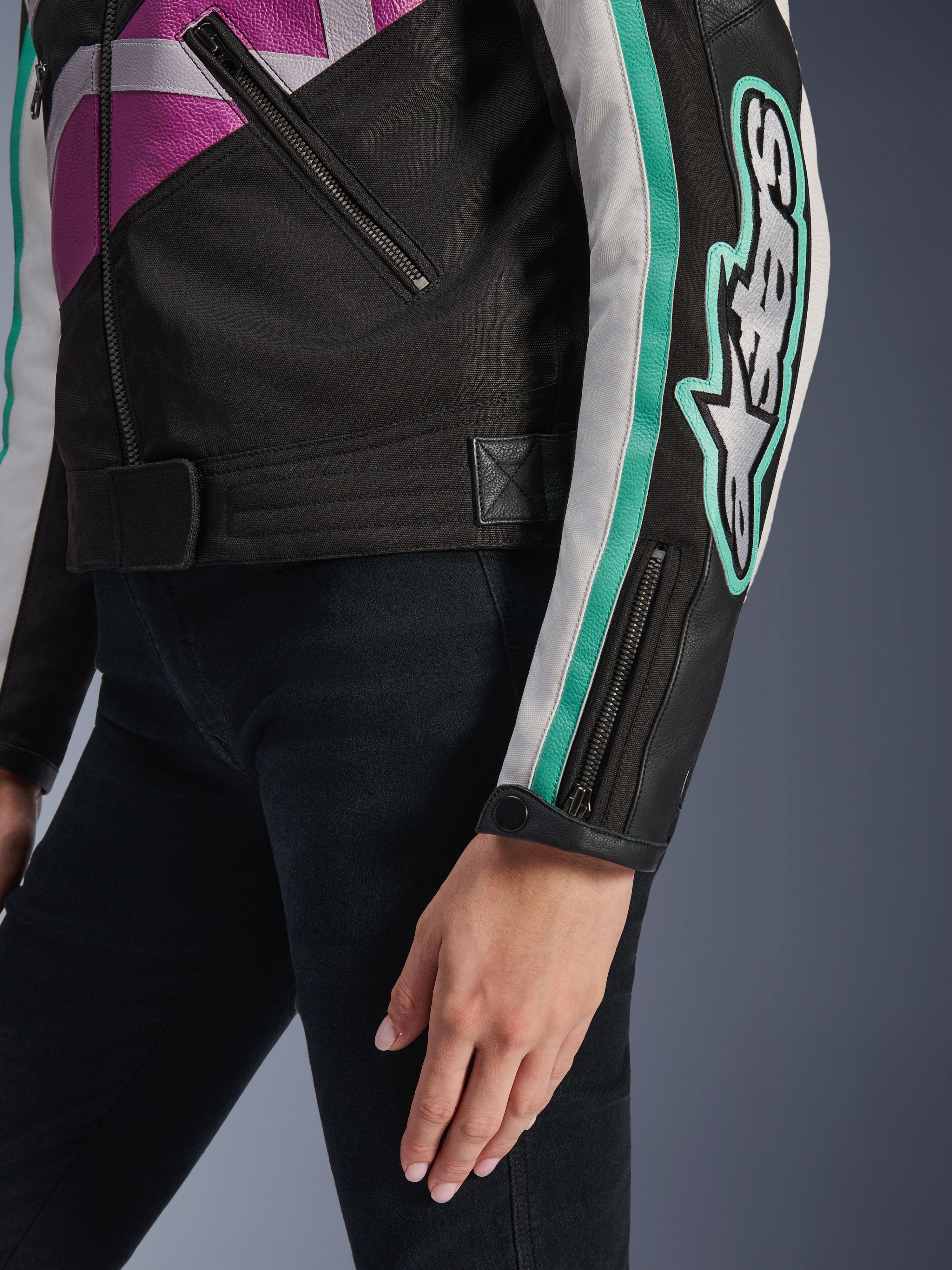 Alpinestars Idles damjacka, närbild av ärmen och sidan av överkroppen, svart med lila, vita och turkosa detaljer, med en framträdande Alpinestars-logotyp på armen, manschett med dragkedja och tryckknapp samt justeringsrem i midjan, vintageinspirerad motorcykelutrustning.