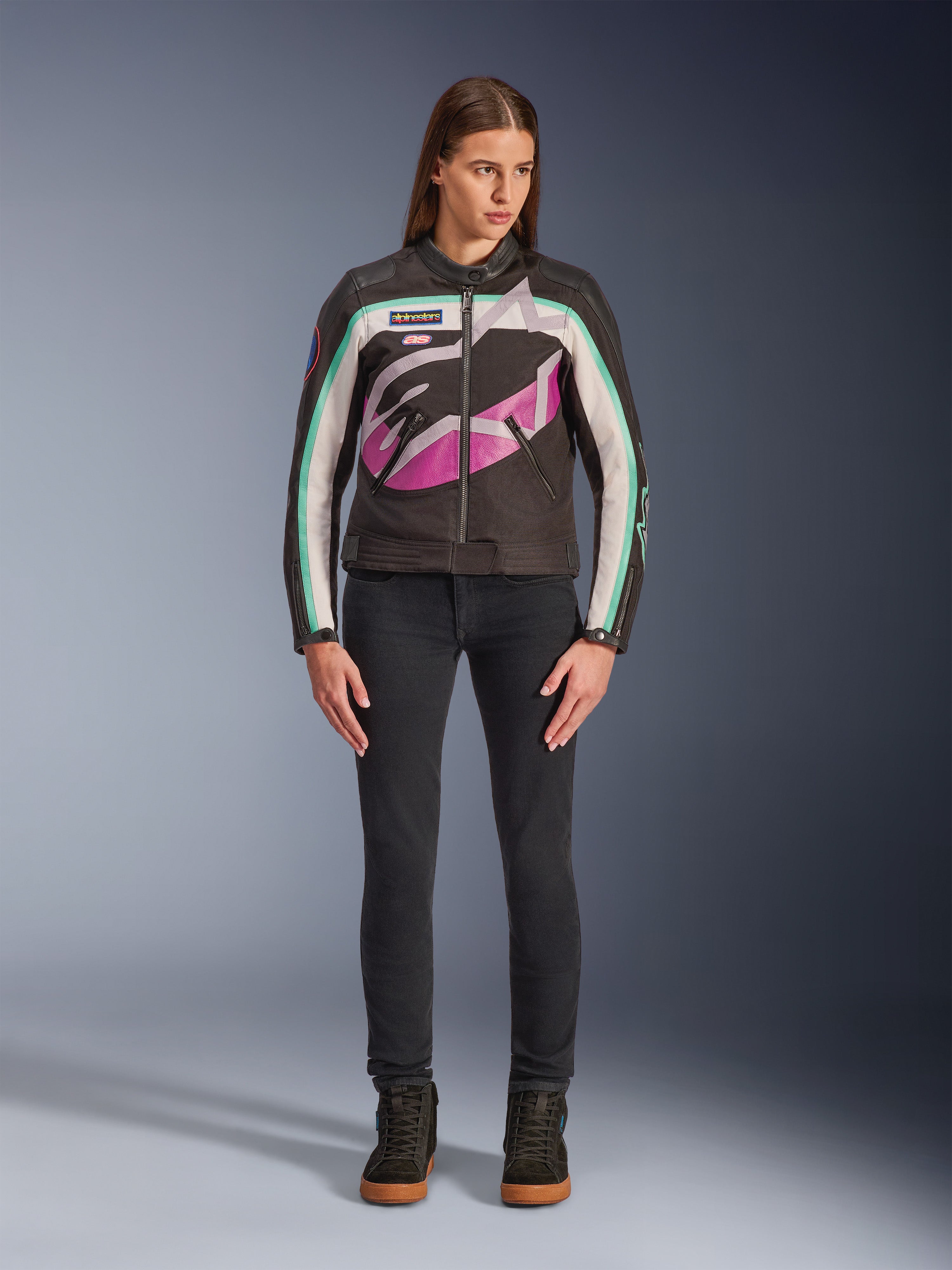Kvinna i Alpinestars Idles damjacka, svart, benvit och lila, vintage racing-inspirerad design med stor omslutande logotyp och blågröna detaljer, stylad med svarta jeans och mc-sneakers, urban motorcykelutrustning