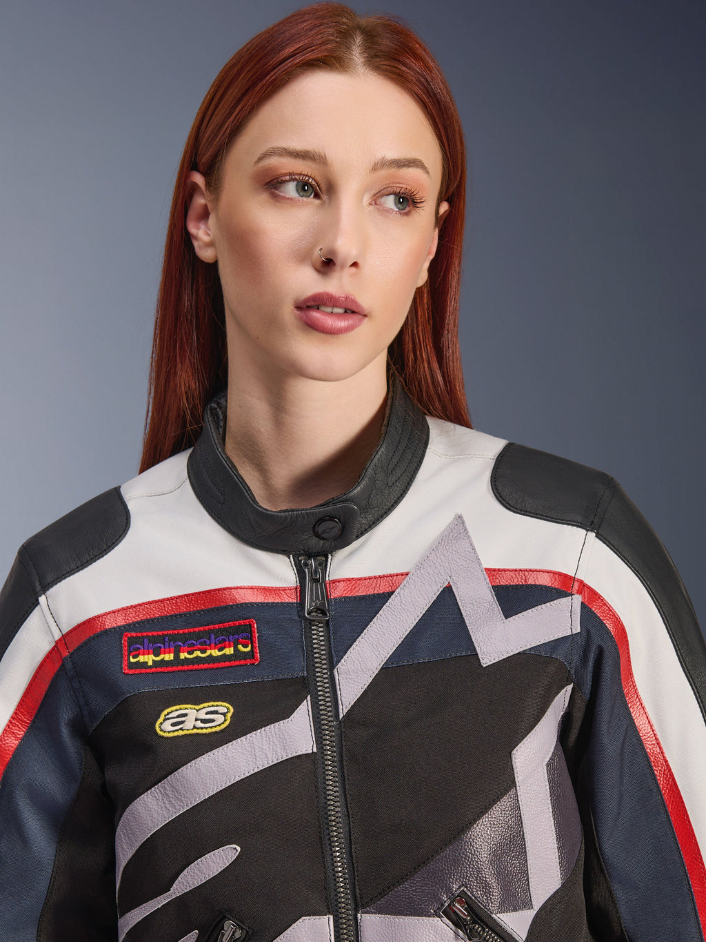 Alpinestars Idles Women’s Jacket, close-up of a female model with red hair, Off White Flame Red and Black colorway, vintage-inspired racing design with logo patches and grey geometric star graphics, urban motorcycle riding gear.