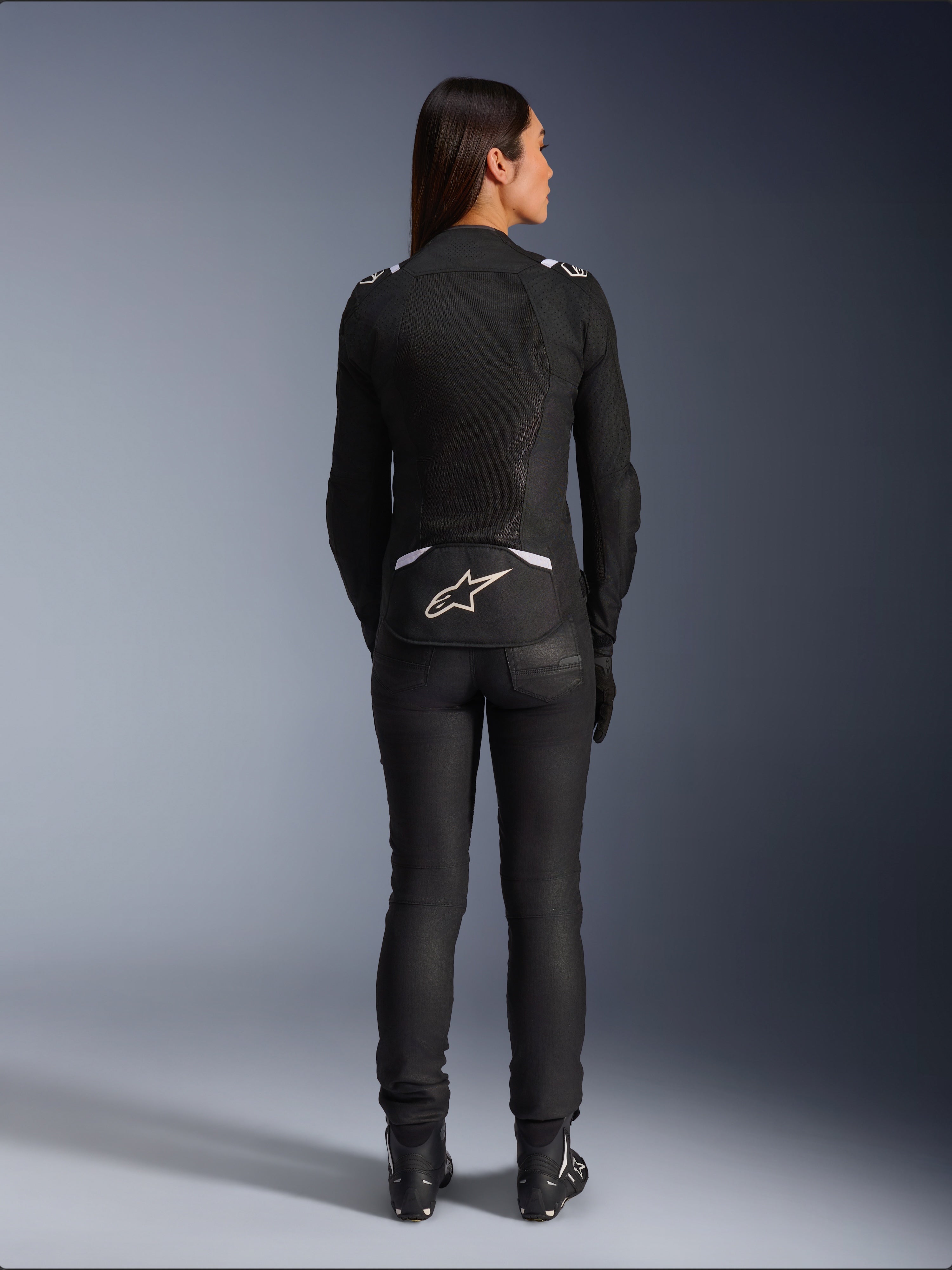 Alpinestars Stella T-SPS Air v2-jacka, motorcykeljacka i textil för dam, svart och vit, vy bakifrån som visar ventilationspaneler i mesh och vit logotyp på nedre delen av ryggen, bärs med svarta mc-byxor och stövlar, urban sportutrustning.