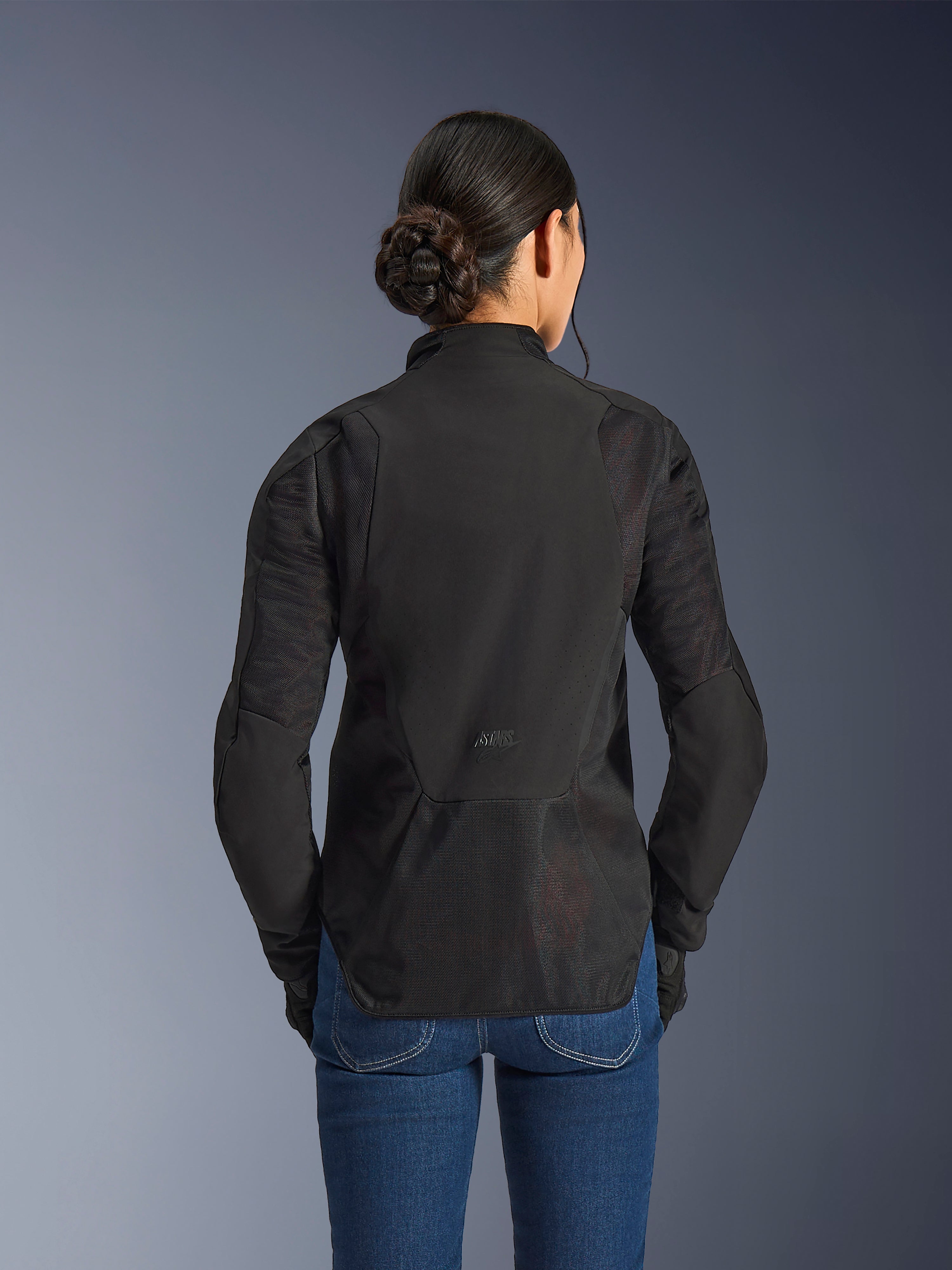 Alpinestars Stella Aeroshell Airflow-jacka för dam, Black Anthracite, vy bakifrån som visar integrerade ventilationspaneler i nät på ryggen och ärmarna, laserperforerat softshell-chassi med förlängd bakre profil, bärs med blå denimjeans.