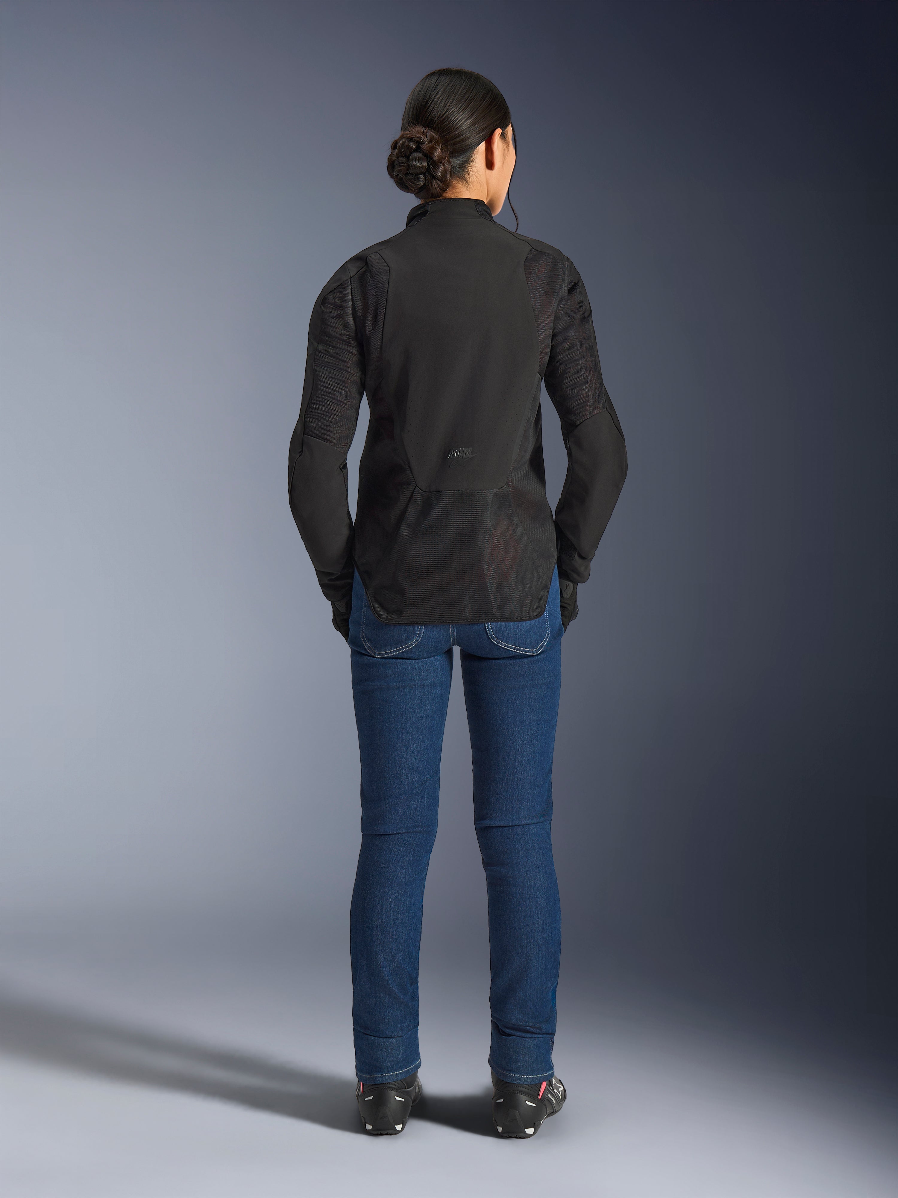 Stella Aeroshell Airflow-jacka för dam, Black Anthracite, vy bakifrån på en modell, med ett lätt softshell-chassi med laserperforerade meshpaneler, förlängd bakre profil för täckning, sportig körpassform, kombinerad med blå jeans.