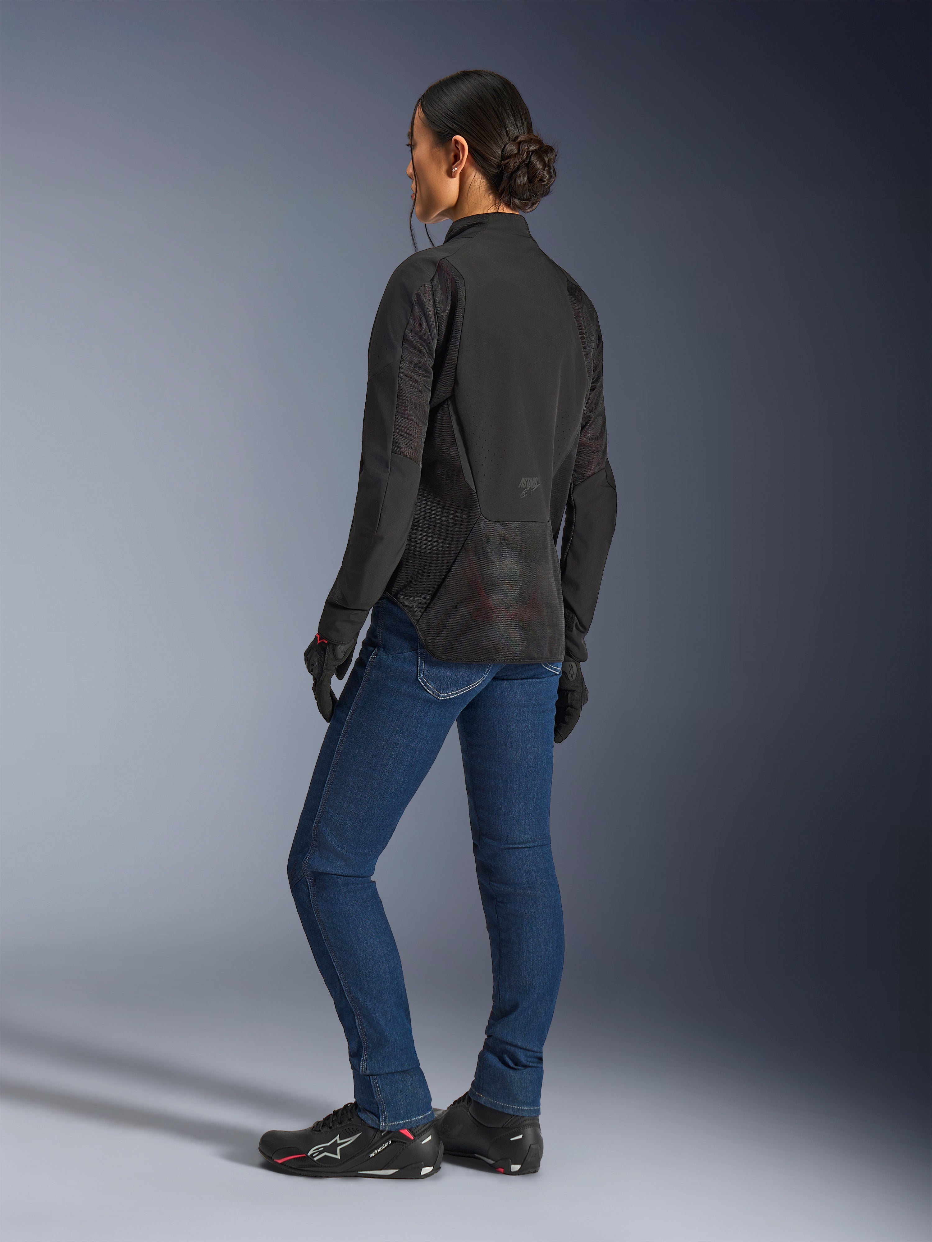 Kvinna som bär Alpinestars Stella Aeroshell Airflow-jacka i Black Anthracite, trekvartsvy bakifrån som visar meshpaneler som andas och förlängd bakre profil, kombinerad med blå MC-jeans i denim och svarta Alpinestars-skor