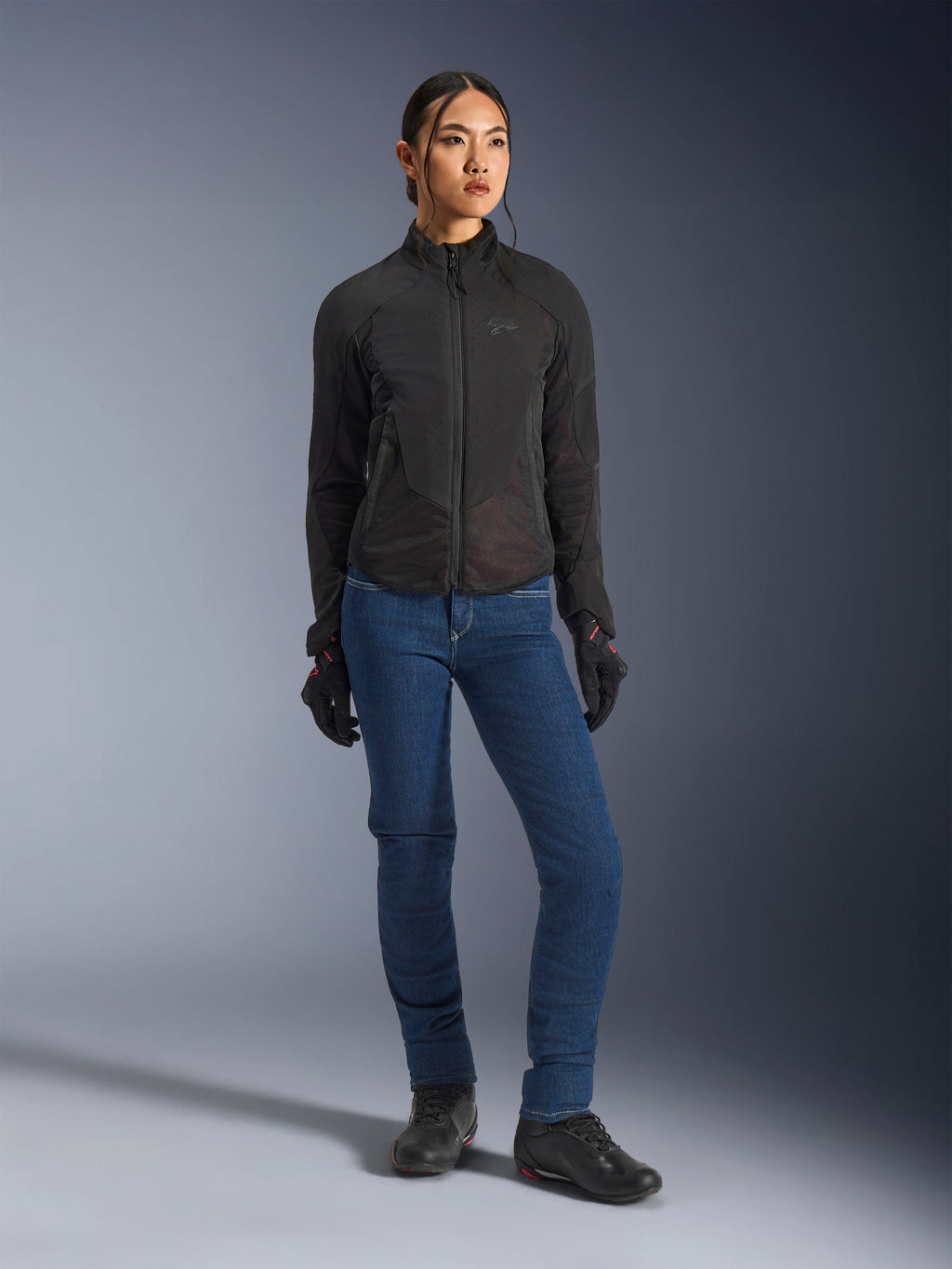 Kvinnlig förare som bär Alpinestars Stella Aeroshell Airflow-jacka i svart, med laserperforerad ventilation och nätpaneler, stylad med blå MC-jeans och svart motorcykelutrustning, modern sportig och urban estetik