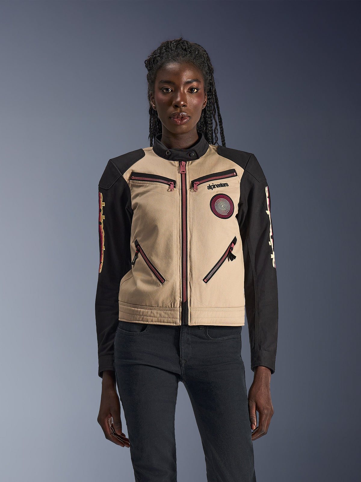 Alpinestars Stella Grace Jacket, Women's Motorcycle Jacket, Aluminum and Black, featuring a workwear-inspired design with pinkish-red zipper accents, a spiral chest patch, and long black sleeves with yellow and red graphics, worn by a model.