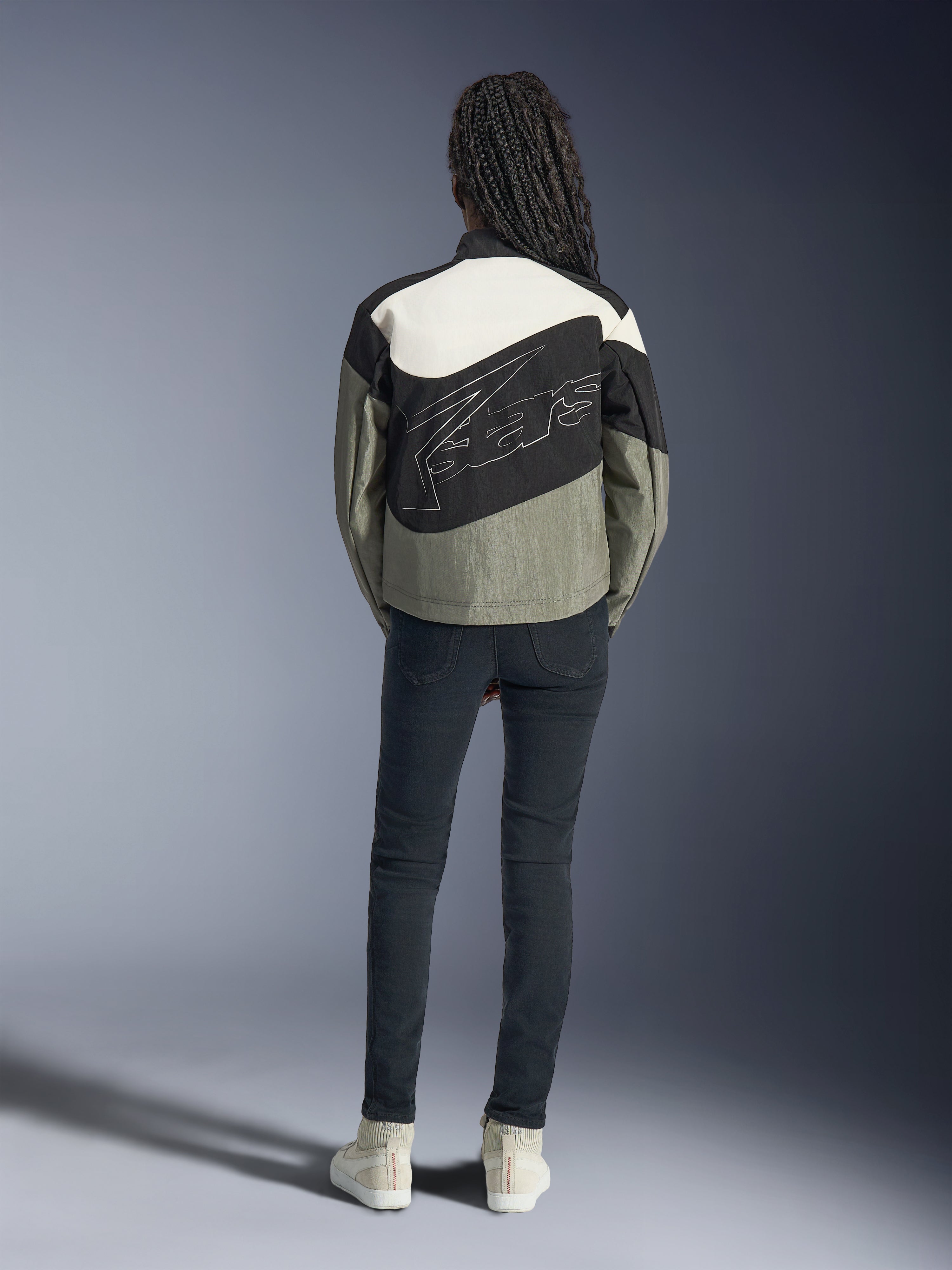 Ryggvy av en modell som bär Alpinestars Stella Moshell-jackan i svart, grått och off-white, med en asymmetrisk design och en stor stiliserad konturlogotyp på ryggen, kombinerat med mörka slim-fit jeans och vita sneakers, urbana streetwear-inspirerade motorcykelkläder för kvinnor.