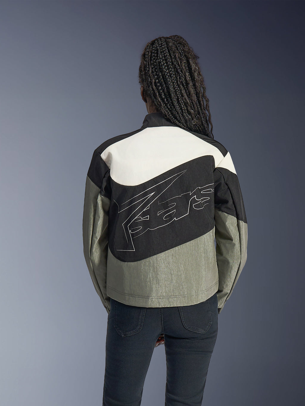 Kvinna sedd bakifrån i Alpinestars Stella Moshell-jacka, svart, grå och off-white urban motorcykeljacka med en stor vit konturerad logotyp på ryggen, matchad med svarta byxor, modern streetwear-inspirerad mc-utrustning.