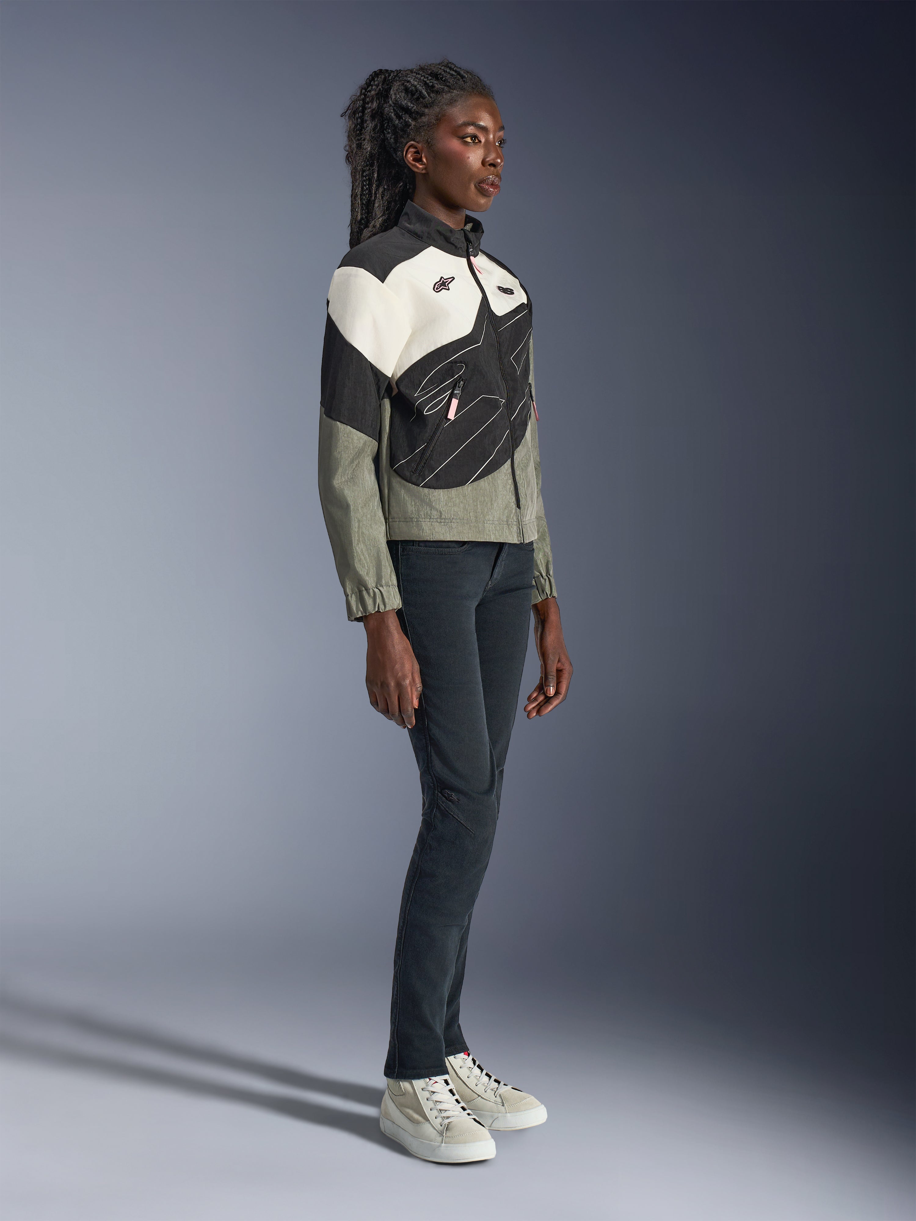 Alpinestars Stella Moshell-jacka, urban motorcykeljacka för dam, grå, svart och off-white med rosa detaljer, helfigursbild av en kvinnlig modell i mörka byxor och sneakers, modern streetwear-inspirerad design för stadskörning