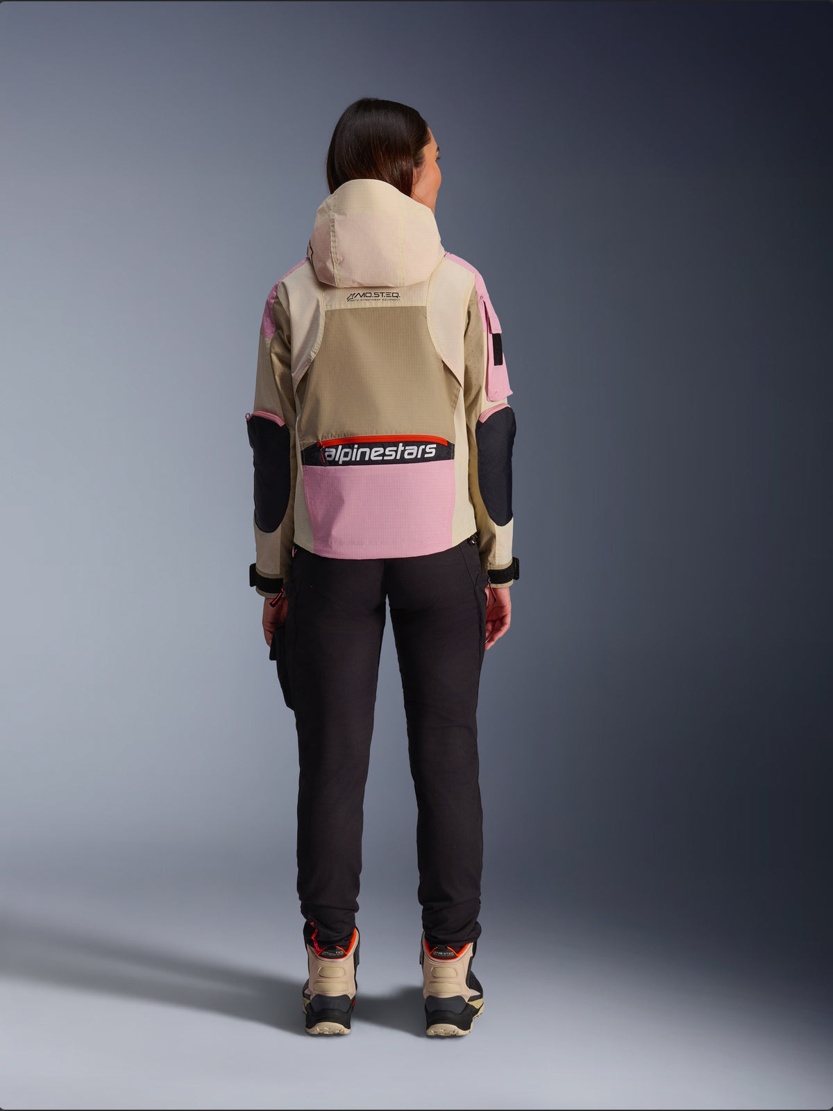 Kvinnlig förare sedd bakifrån iförd Alpinestars Molly Air-jacka i Sand Pink Almond, med beige huva och rosa paneler med svarta detaljer, matchad med svarta mc-byxor och beige motorcykelstövlar, urban samtida motorcykelutrustning