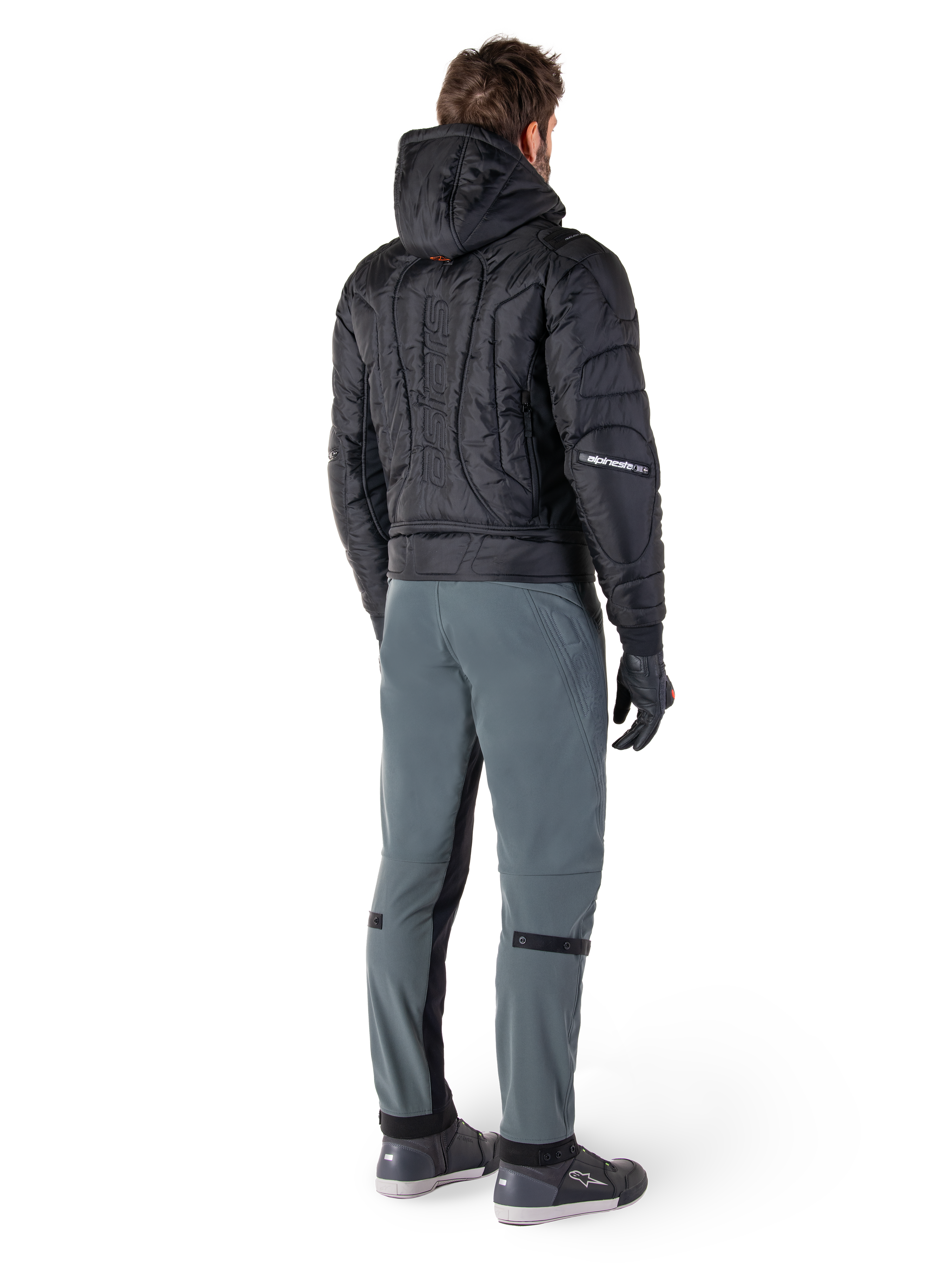 Alpinestars Alden-byxor, urbana motorcykelbyxor, Urban Gray, baksida av förare i svart quiltad jacka med huva och motorcykelsneakers, har en smal passform under knät med svarta stretchpaneler och knäjusteringar, modern streetwear-inspirerad design.