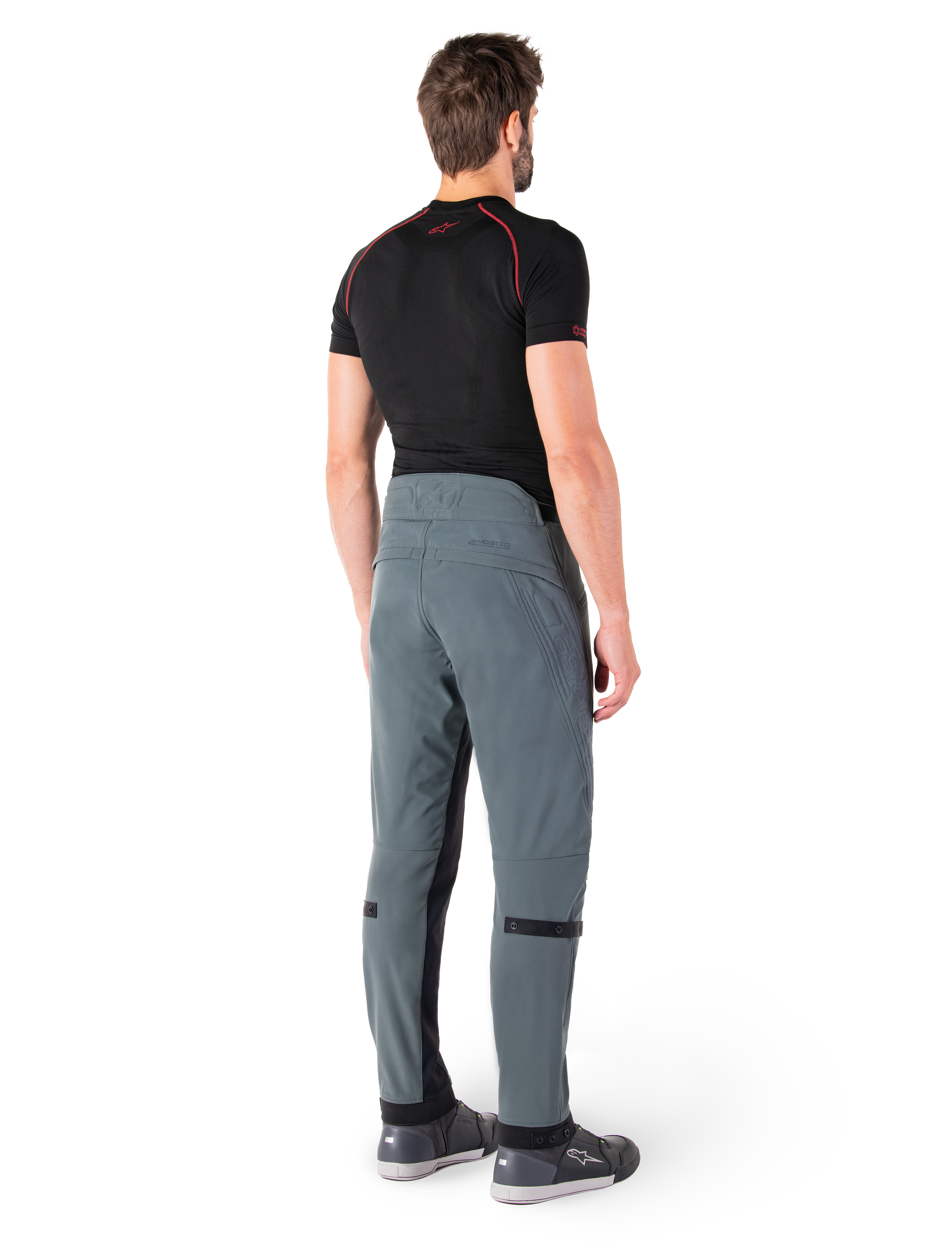 Alpinestars Alden byxor, Urban Gray, sedda bakifrån, tekniska motorcykelbyxor i textil med kviltade detaljer på den övre bakre linningen och benen, kombinerade med ett svart baslager och grå motorskor, modern urban streetwear-stil.