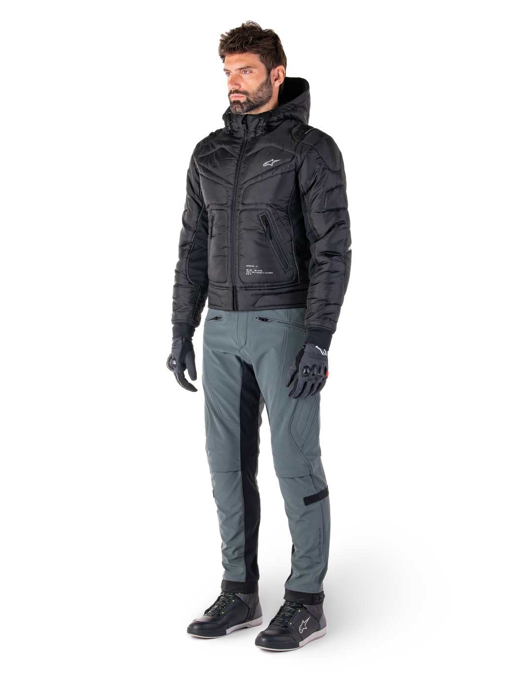 Alpinestars Alden-byxor i Urban Gray, bärs av en modell med en svart kviltad jacka med huva, handskar och motorskor, med kviltade detaljer på sidorna och svarta paneler på innerbenen, streetwear-inspirerad urban motorcykelutrustning.