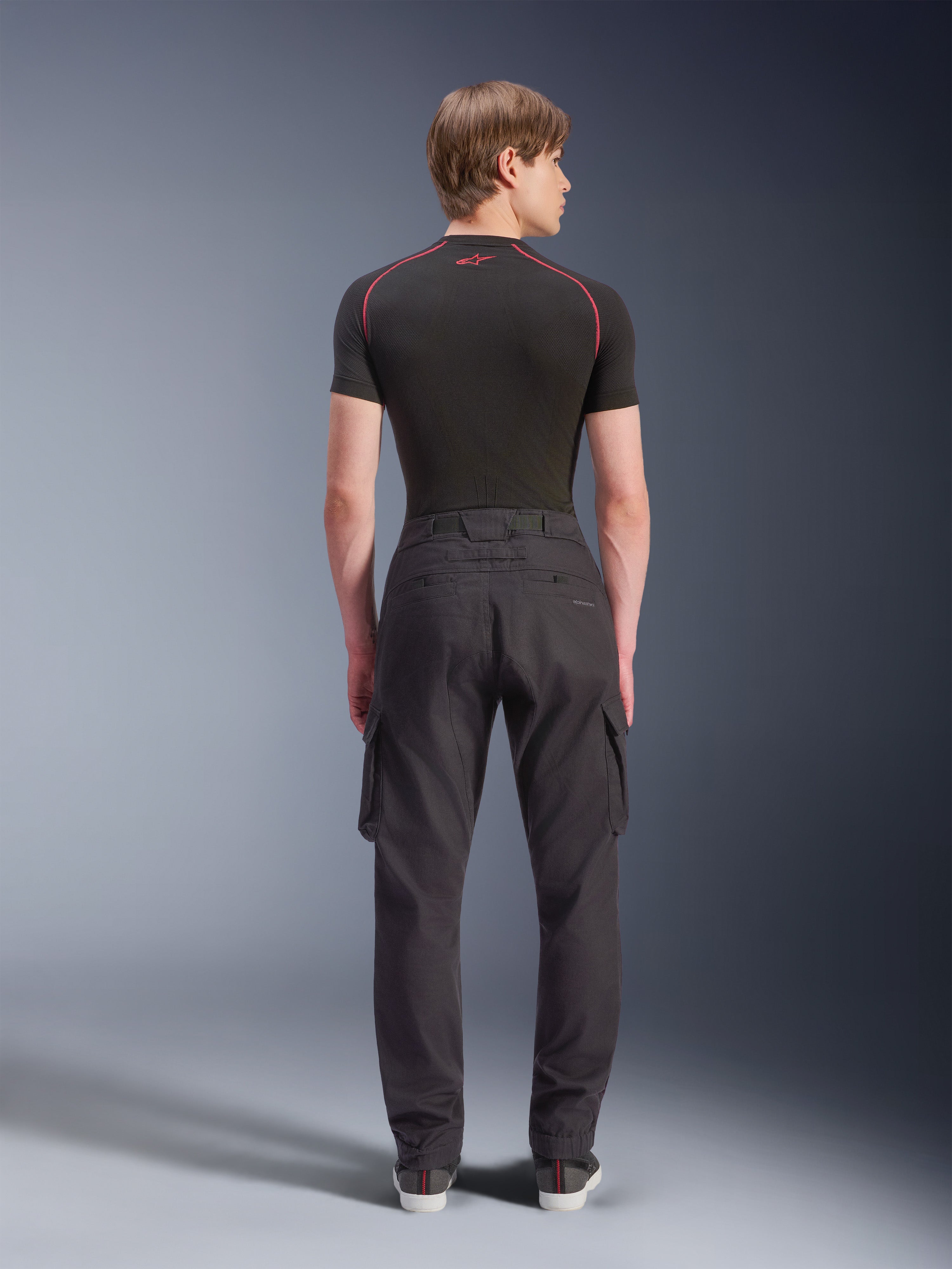 Alpinestars Flex-AST Cargo Pants - Short, svarta urbana motorcykelbyxor sedda bakifrån, med cargofickor på sidorna och elastiska benslut, bärs med ett svart kortärmat underställ med röda kontrastsömmar