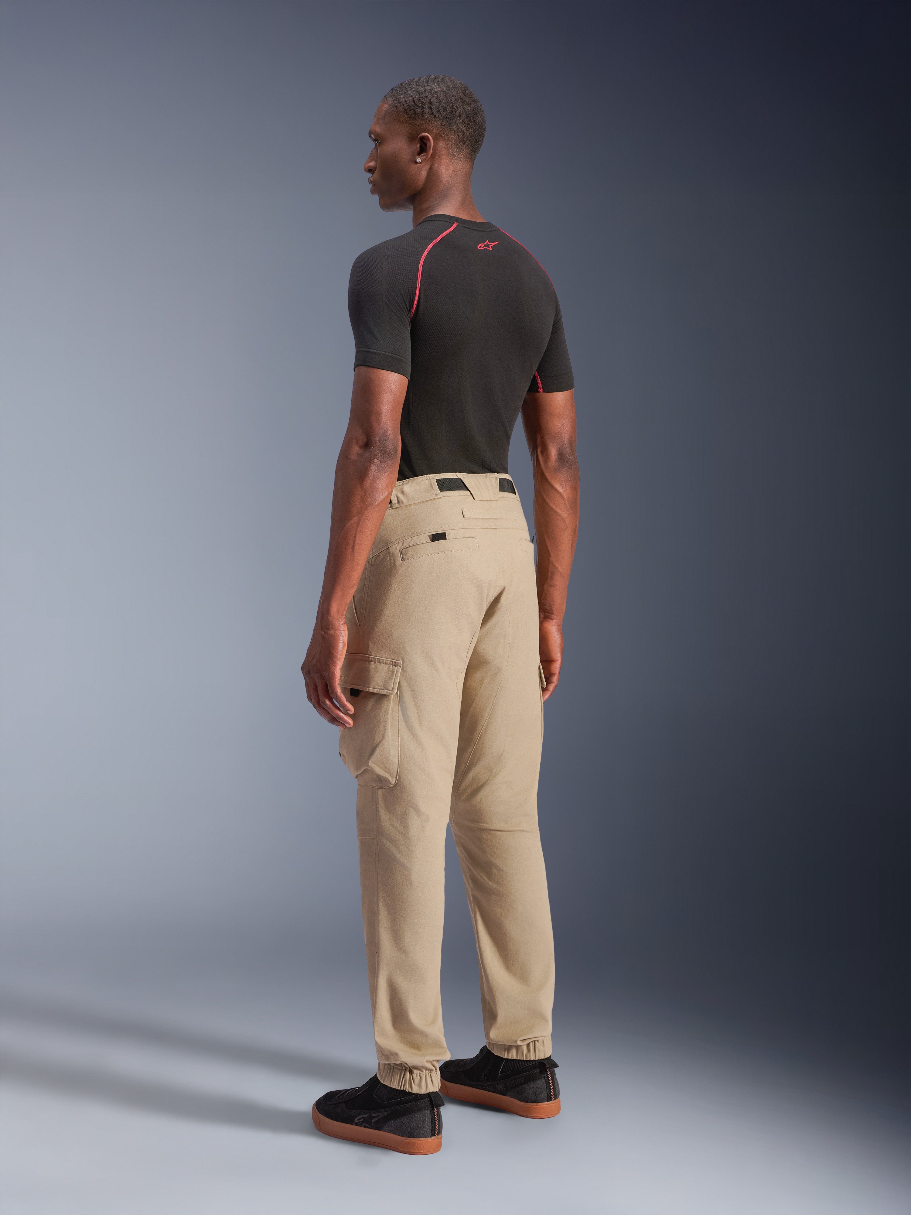 Alpinestars Flex-AST Cargo Pants, urbana motorcykelbyxor, ljusbeige, visas från en vinkel snett bakifrån, med funktionella cargofickor och elastiska benslut, matchas med en svart kortärmad tröja och svarta sneakers.