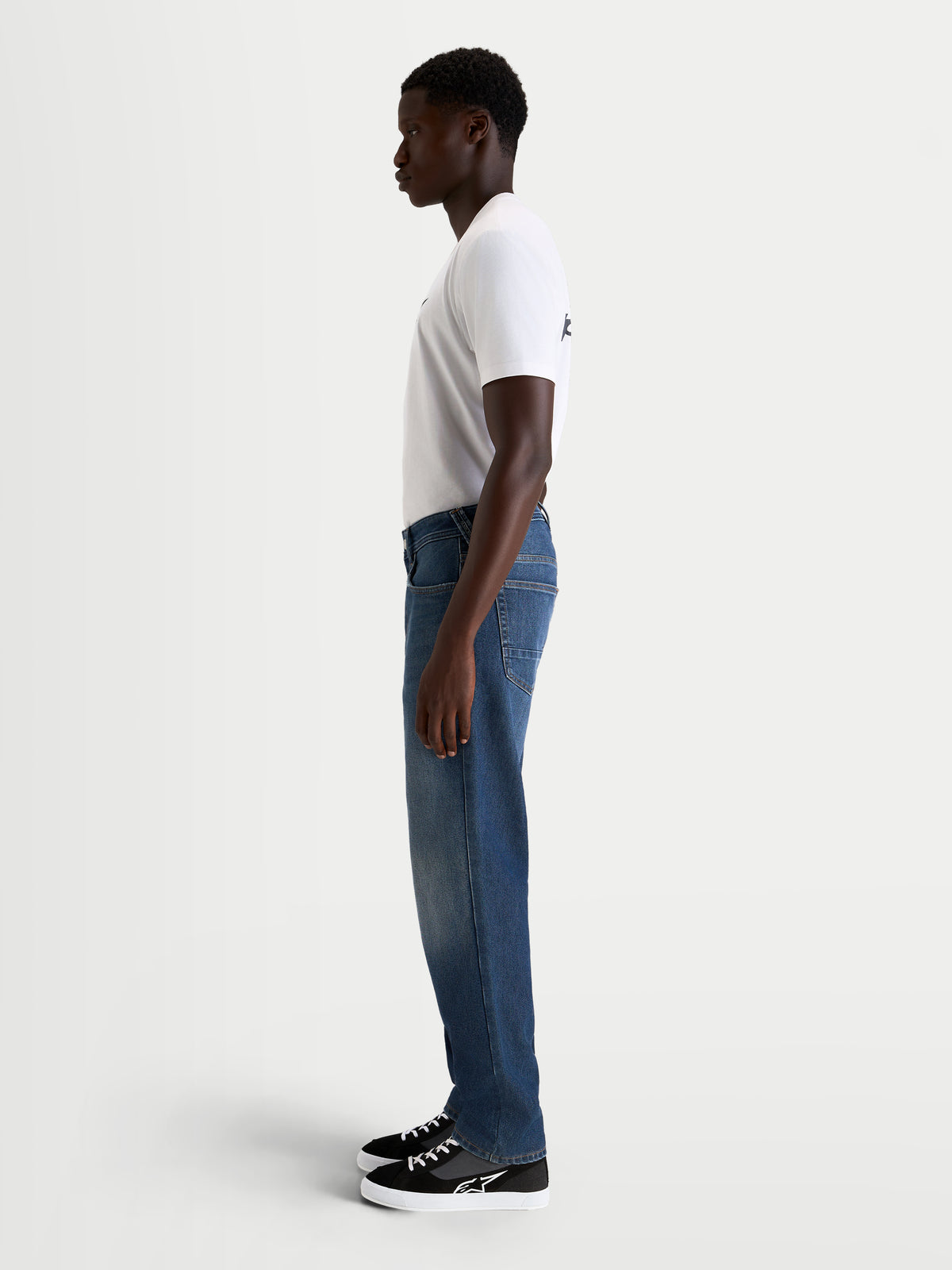 Alpinestars Flex-AST 8 denim byxor, urbana motorcykeljeans för herr i mellanblått, sidoprofil på en modell iklädd vit t-shirt och vardagliga körskor, AA-klassad enlagersdenim med stretchkonstruktion