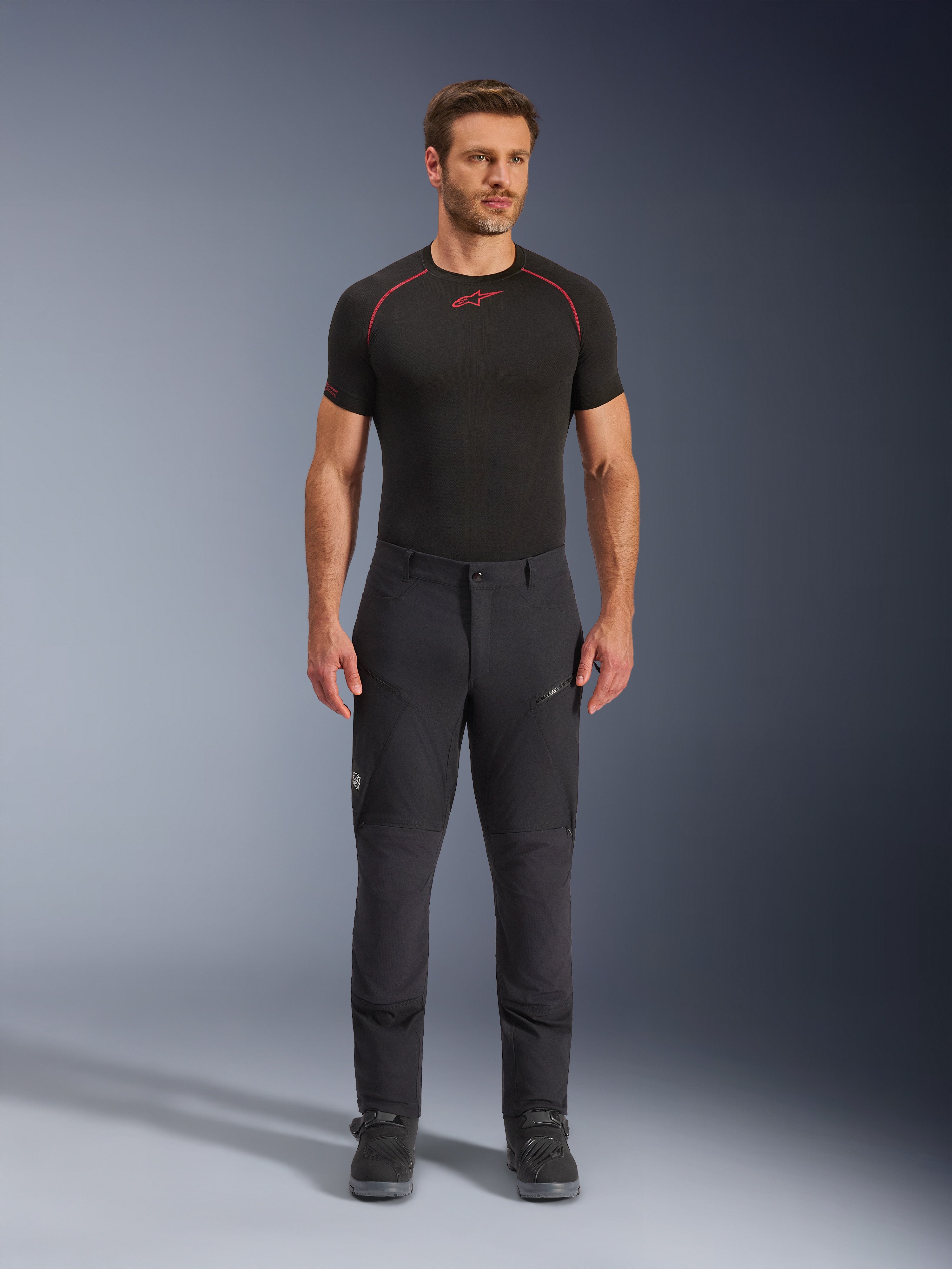 Alpinestars Flex-AST Explorer Pants, svarta äventyrs- och touringbyxor för motorcykel, man som bär åtsittande textilbyxor med cargofickor med dragkedja och förstärkta paneler, matchade med ett svart underställ och motorcykelstövlar, slitstarkt skydd för äventyrskörning