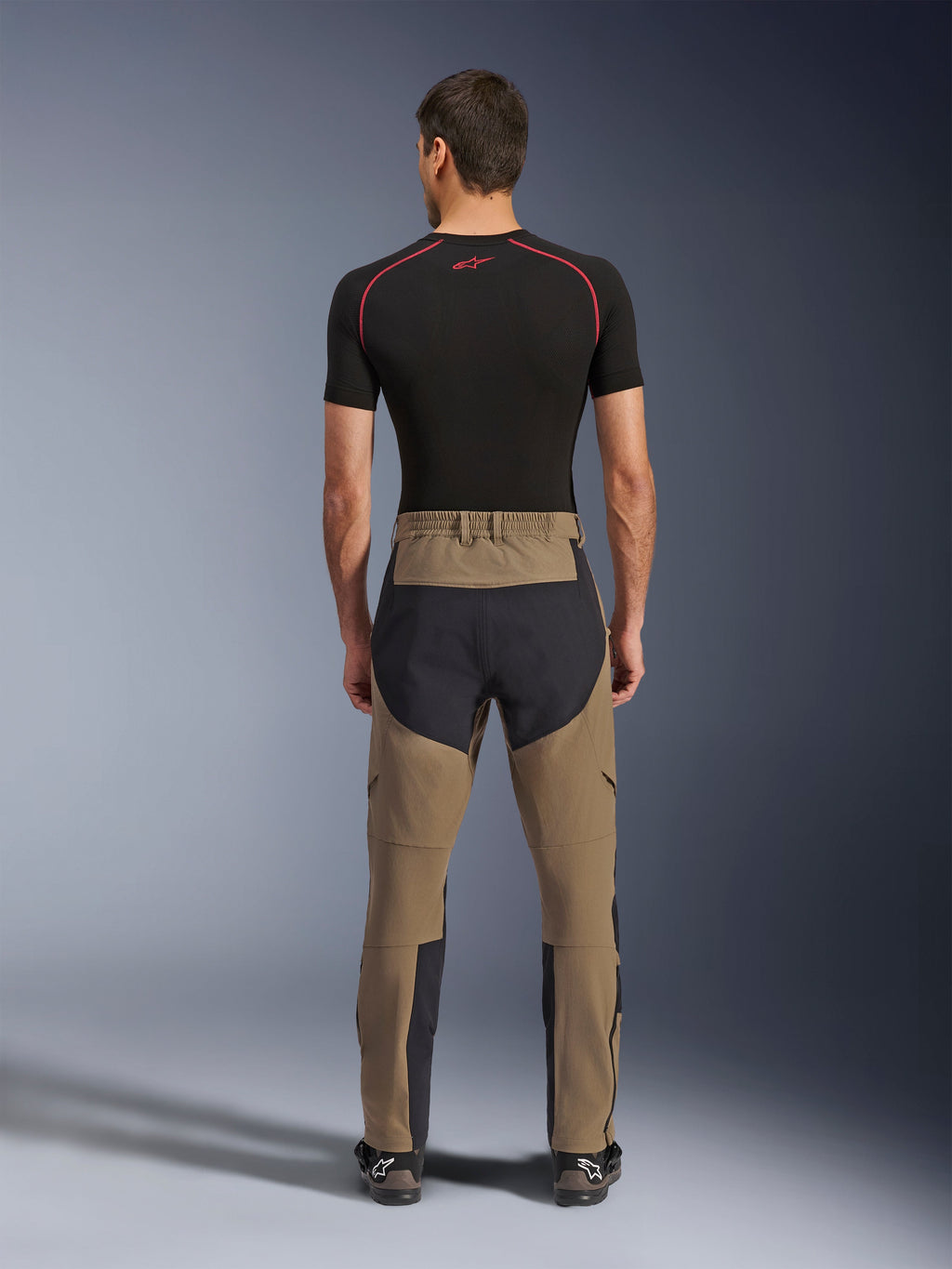 Alpinestars Flex-AST Explorer-byxor, Walnut Black, sedda bakifrån på modell, med förstärkt svart säte och brunt stretchmaterial, kombinerade med svart baslager och tekniska motorcykelstövlar, slitstark utrustning för äventyrskörning