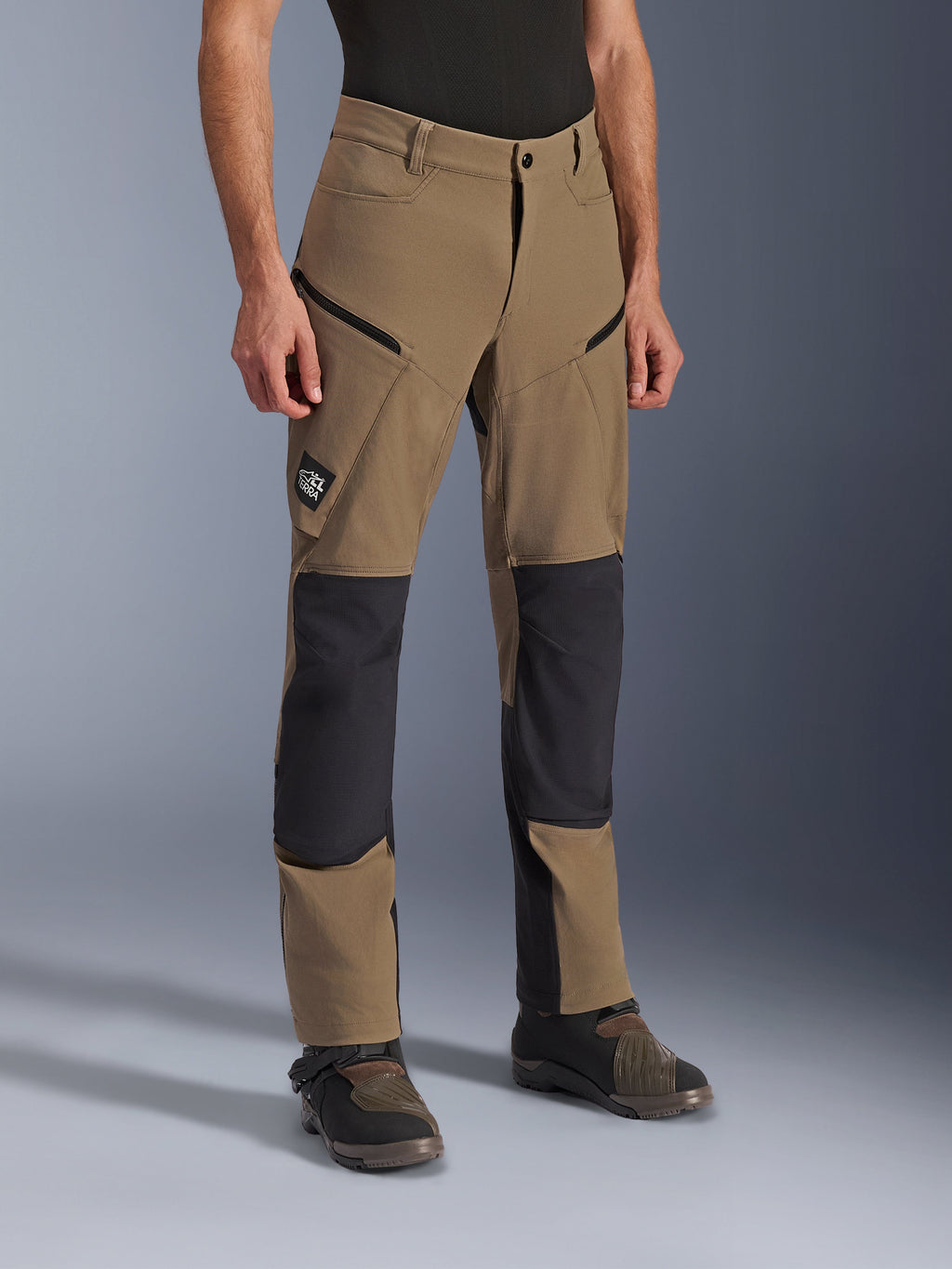 Alpinestars Flex-AST Explorer-byxor, äventyrsmotorcykelbyxor i Walnut Black, med brunt textilmaterial och svarta förstärkta paneler vid knän och säte, cargofickor med dragkedja och All Terra-logotyp, visas tillsammans med tekniska körstövlar.