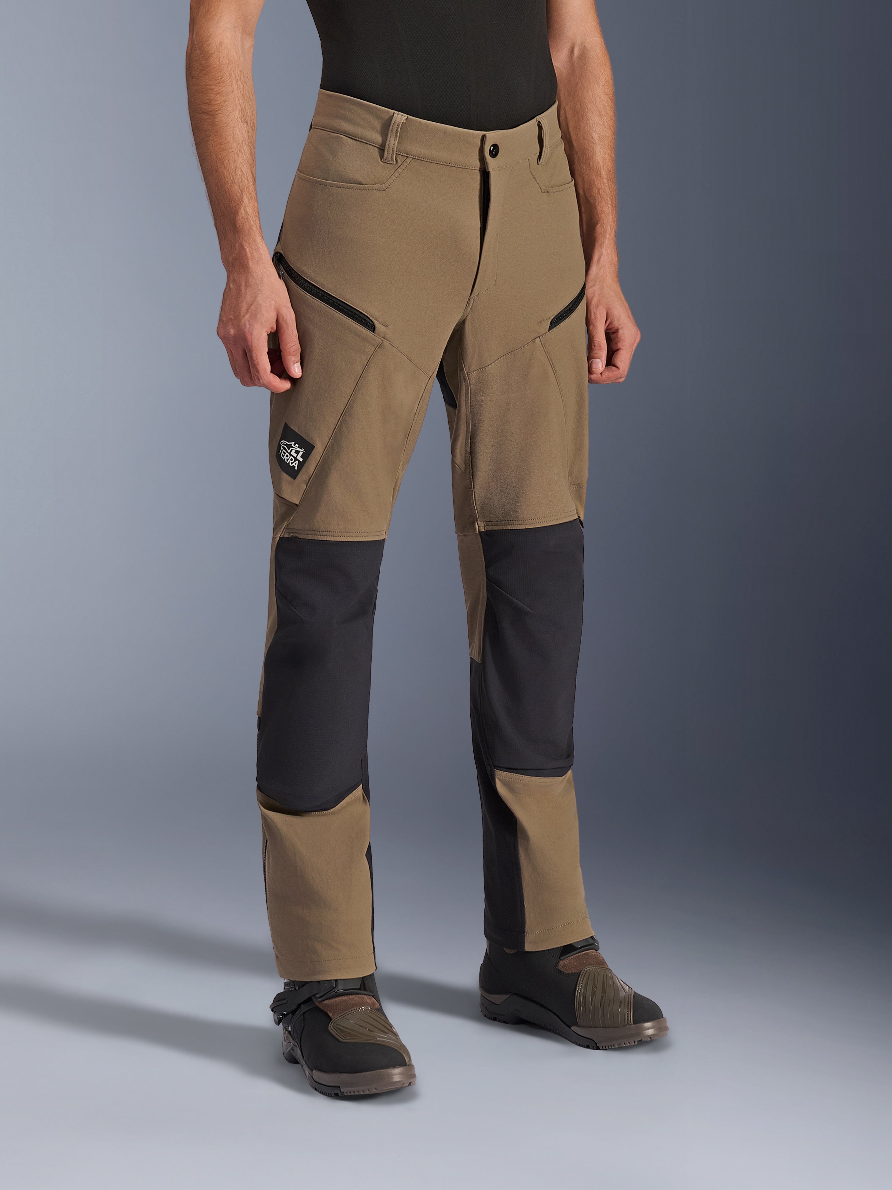 Alpinestars Flex-AST Explorer-byxor, äventyrsmotorcykelbyxor i Walnut Black, med brunt textilmaterial och svarta förstärkta paneler vid knän och säte, cargofickor med dragkedja och All Terra-logotyp, visas tillsammans med tekniska körstövlar.